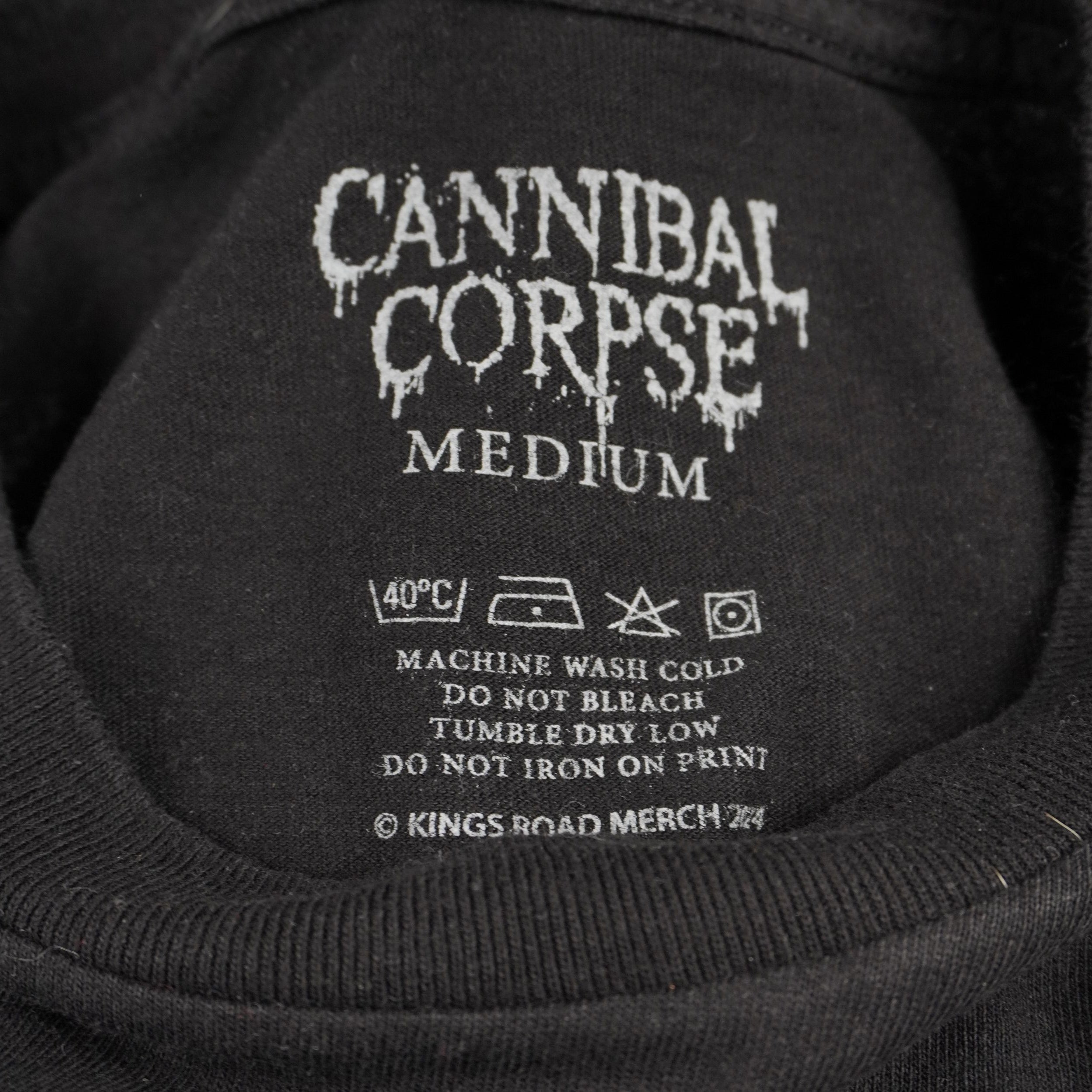 Primo piano dell'interno del colletto della t-shirt che mostra il logo Cannibal Corpse e le informazioni sulla taglia Medium (M) stampate direttamente sul tessuto per un design senza etichetta.