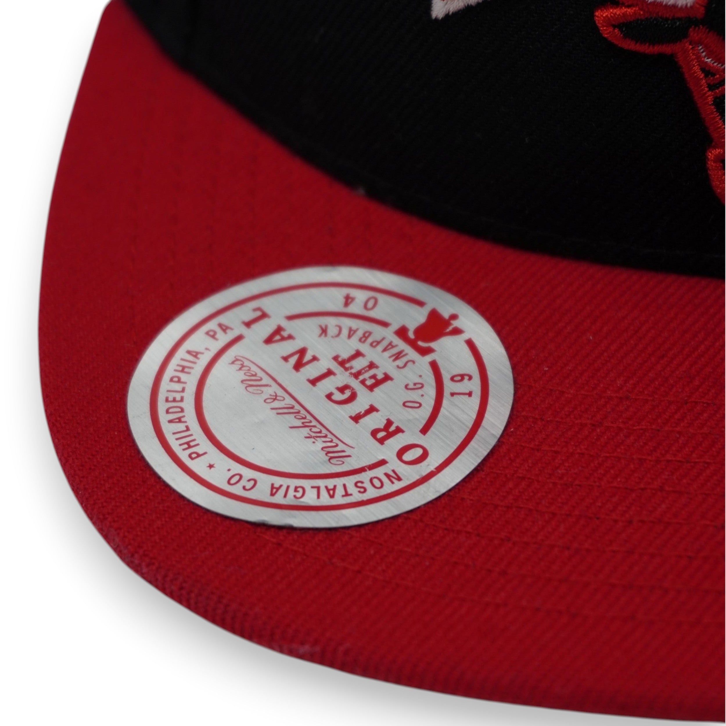  Dettaglio dell'adesivo circolare Mitchell & Ness "Original Fit" sulla visiera rossa piatta del cappello NBA.