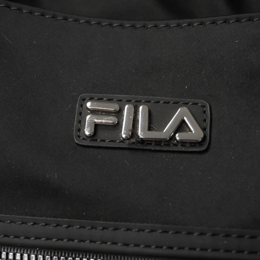  Primo piano della placchetta rettangolare in metallo lucido con marchio FILA a rilievo montata su supporto gommato nero.