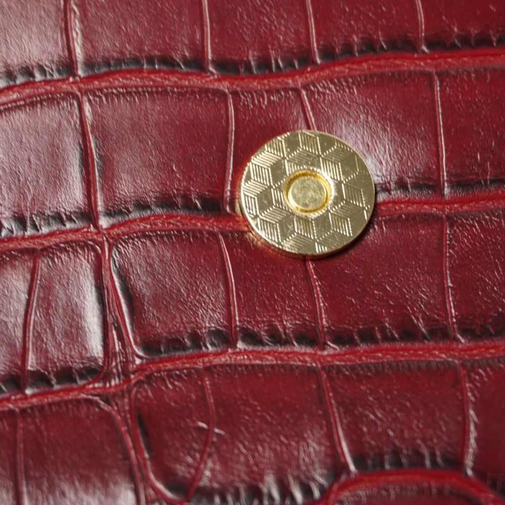 Primo piano del bottone magnetico circolare dorato con lavorazione geometrica zigrinata applicato sulla pelle stampa cocco bordeaux della borsa La Martina.