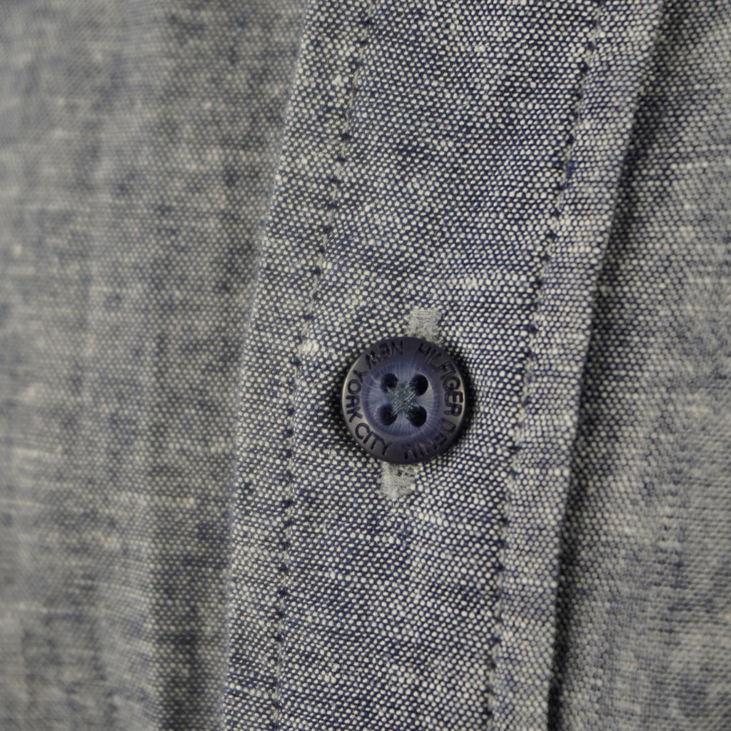 Dettaglio di un bottone blu logato cucito su tessuto grigio in misto lino, particolare della manifattura Hilfiger Denim.