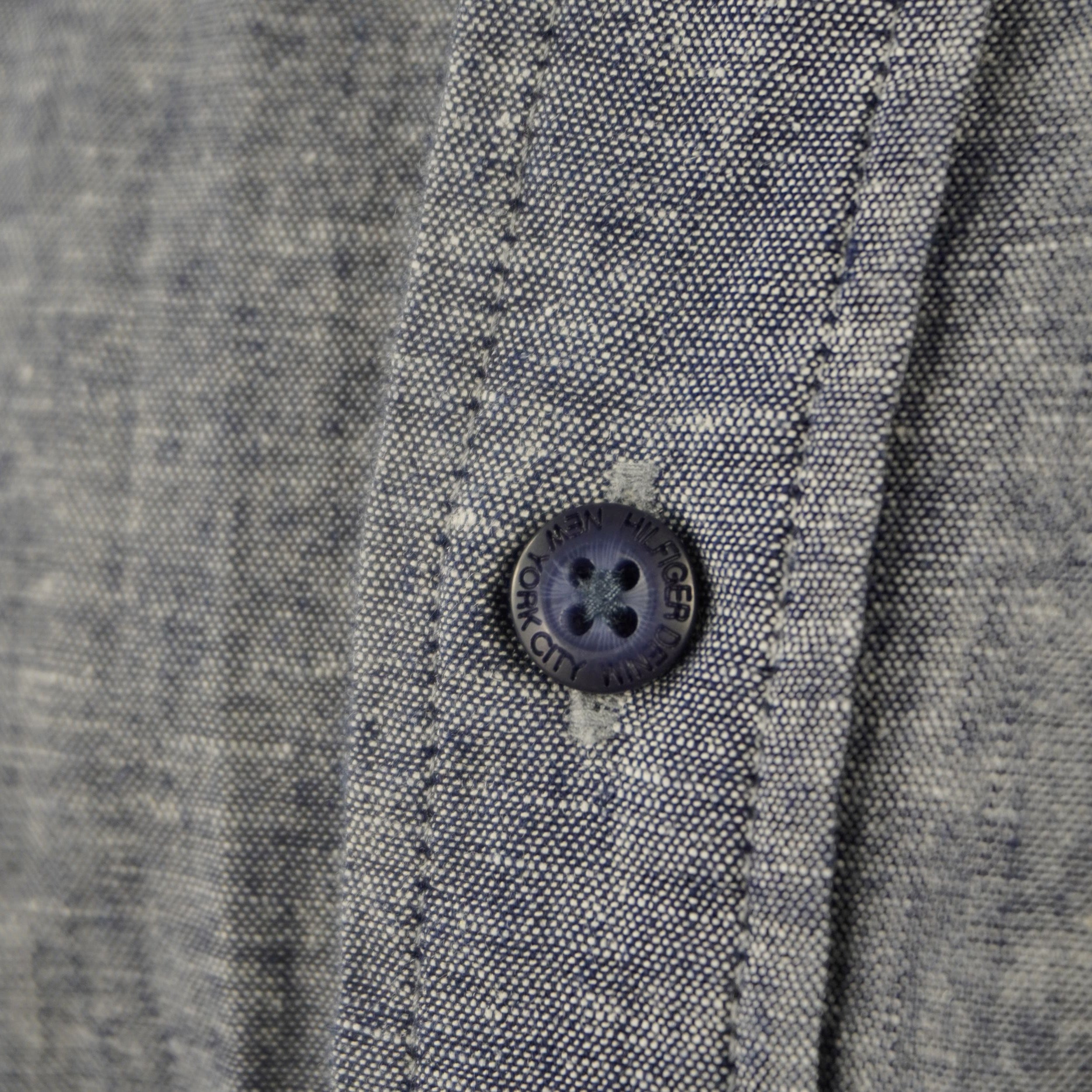 Dettaglio di un bottone blu logato cucito su tessuto grigio in misto lino, particolare della manifattura Hilfiger Denim.