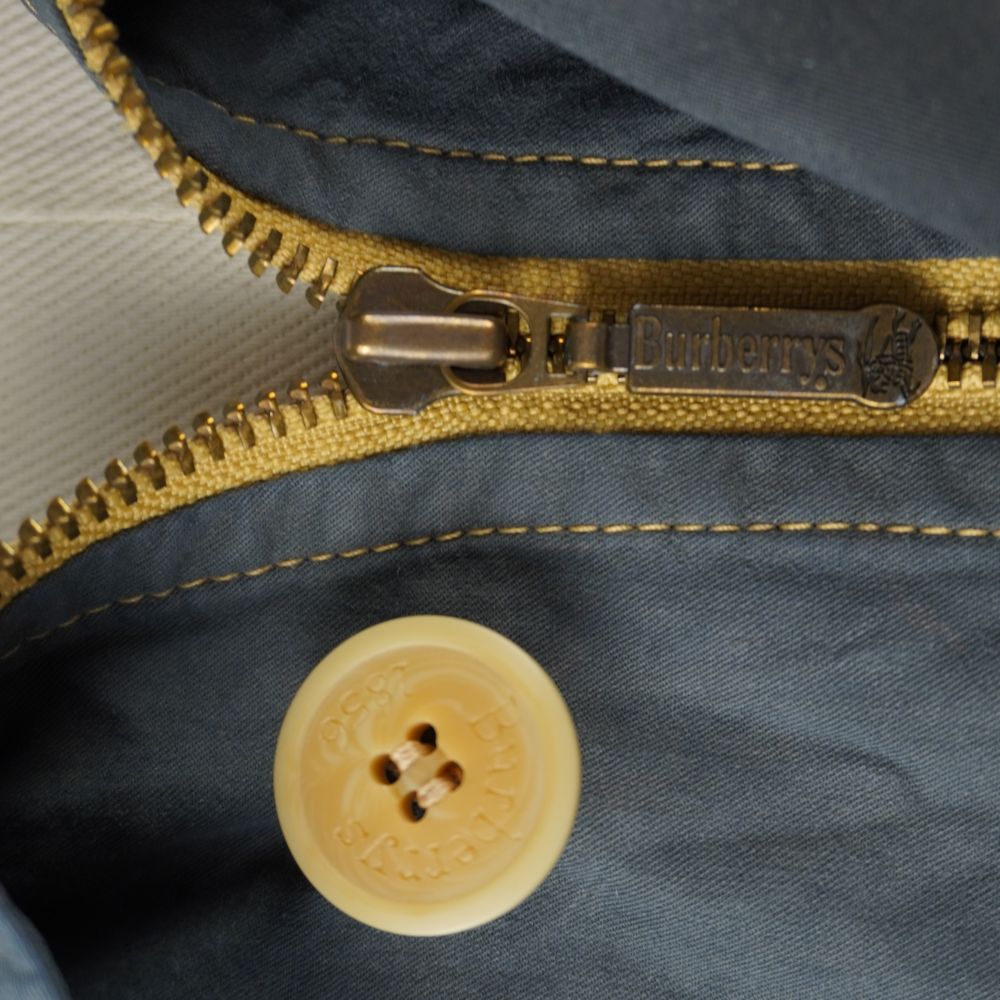 Dettaglio ravvicinato della cerniera lampo in ottone dorato e del bottone inciso Burberrys 1856 sulla patta frontale.