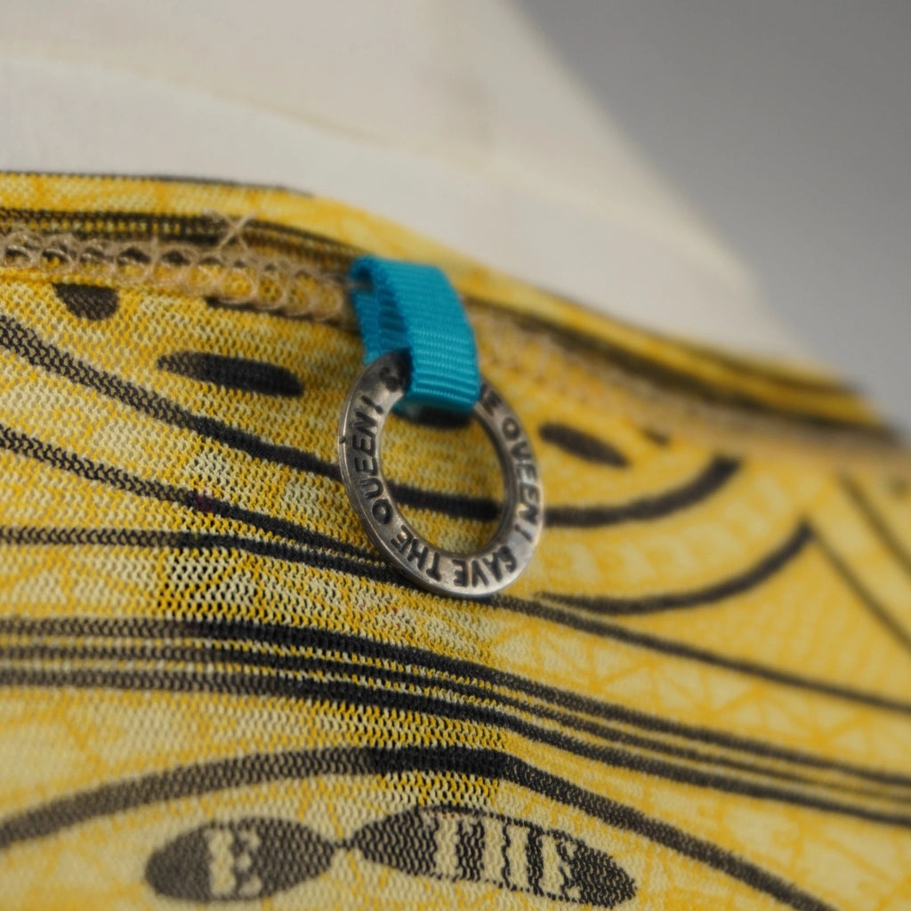 Macro del piccolo ciondolo circolare in metallo inciso con il brand Save The Queen! sul retro del collo.