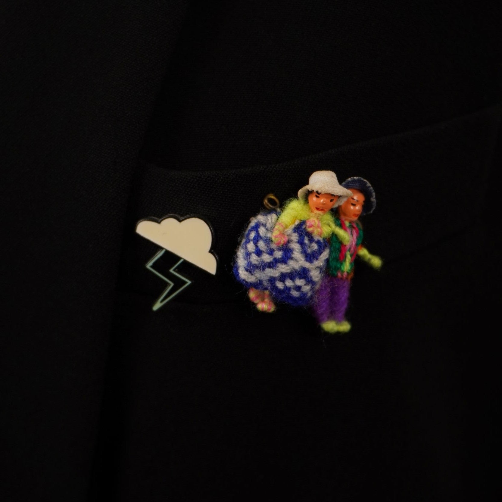 Primo piano sul taschino del blazer nero che mostra due piccole figure tradizionali colorate realizzate all'uncinetto applicate sul tessuto.