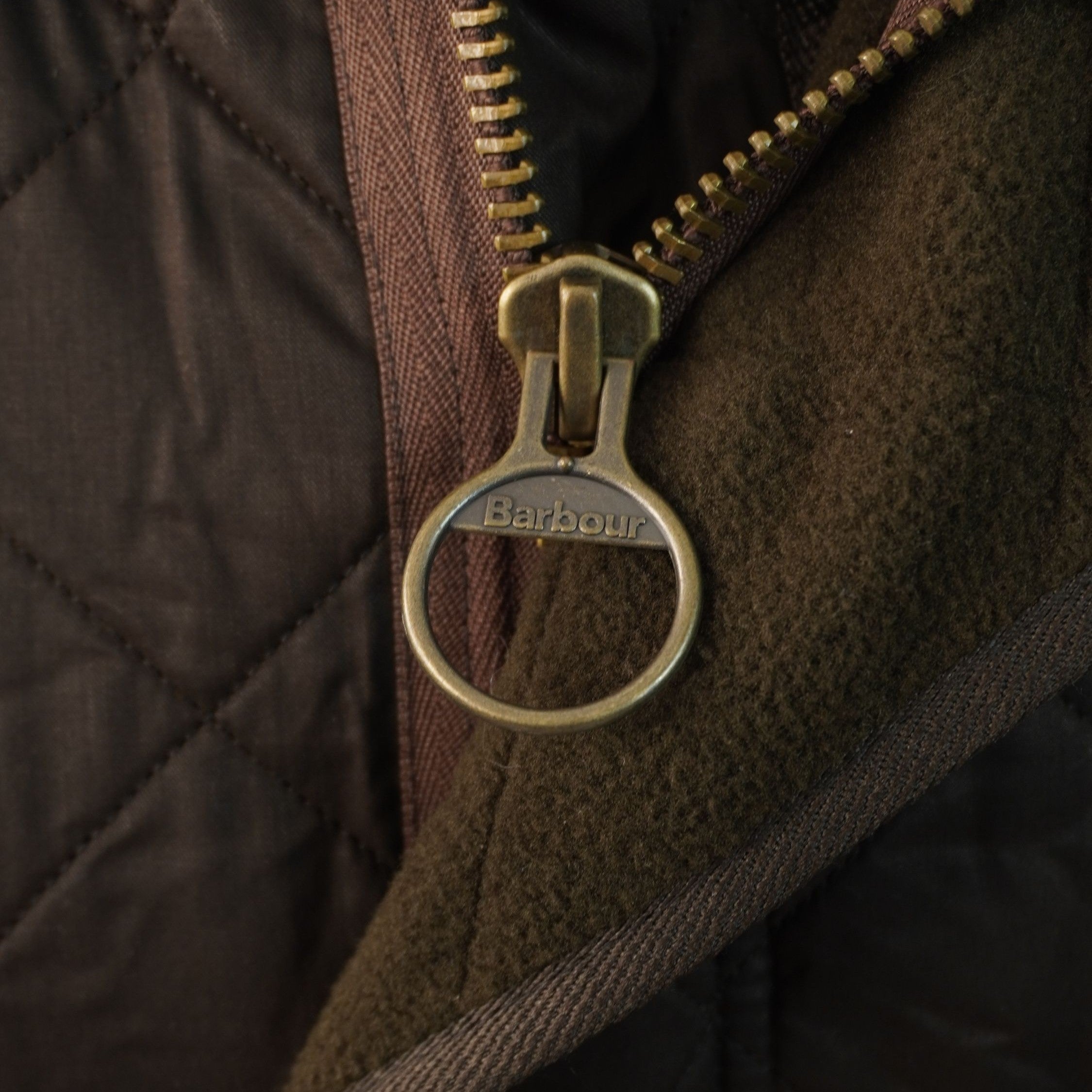 Dettaglio ravvicinato dell'iconica cerniera metallica dorata con cursore ad anello e del colletto in velluto marrone Barbour.