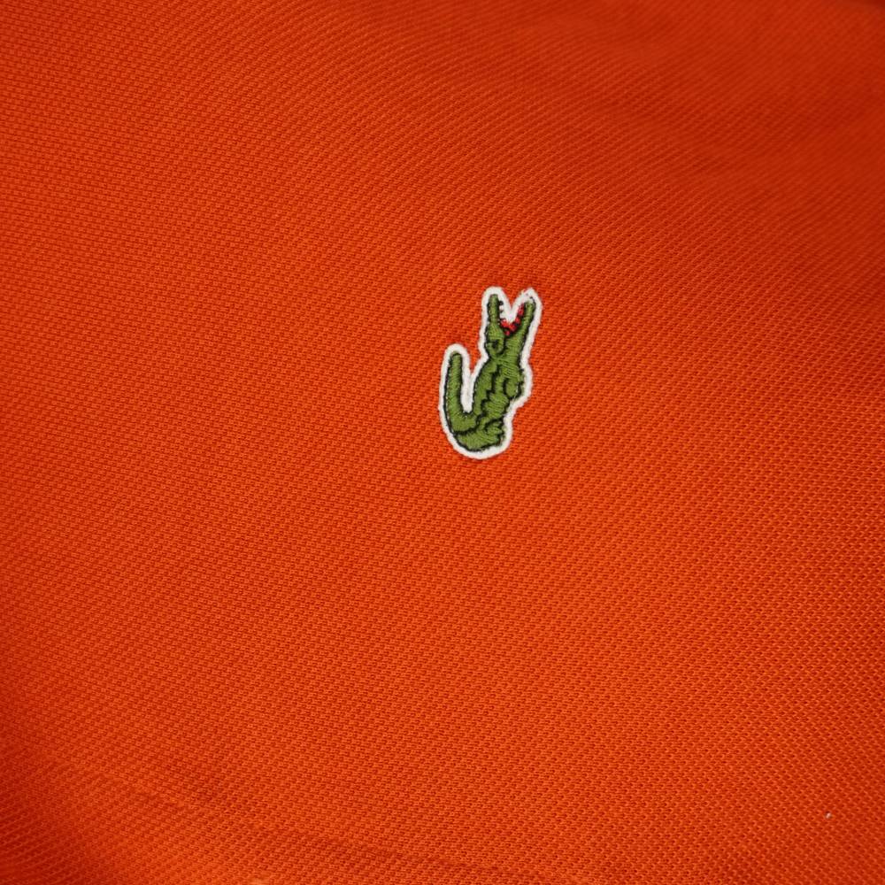 Dettaglio macro del logo coccodrillo verde Lacoste ricamato su tessuto piqué arancione