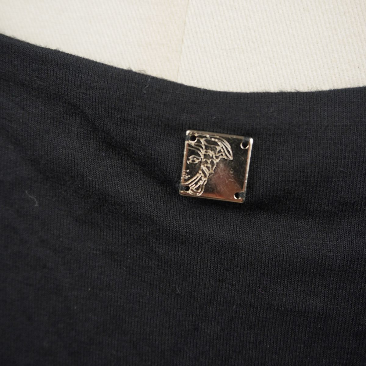 Primo piano ravvicinato della targhetta quadrata in metallo argentato con inciso a sbalzo il logo Medusa di Versace, applicata sul retro del collo.