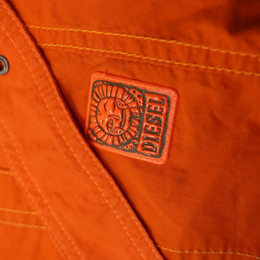 Primo piano della patch logo originale Diesel in gomma 3D con l'iconico sole antropomorfo su tessuto arancione.