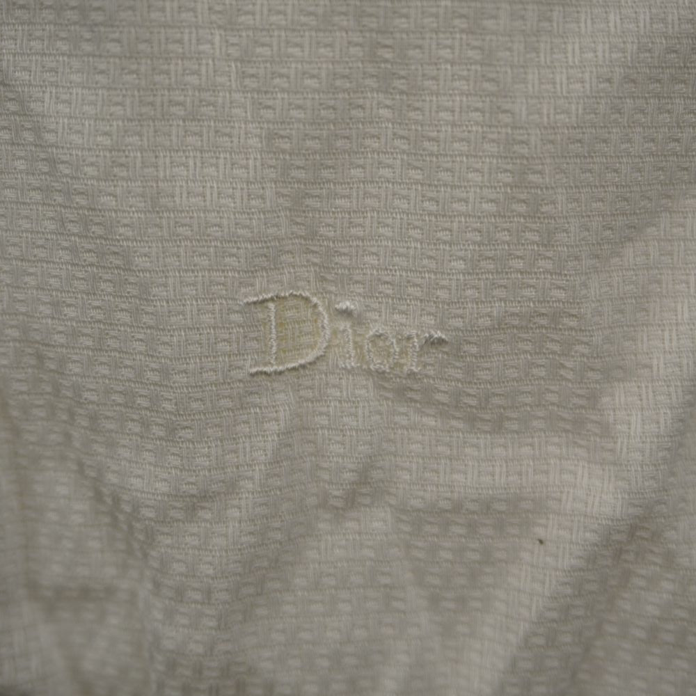 Primo piano del logo Christian Dior ricamato tono su tono sul tessuto jacquard geometrico bianco crema della camicia.
