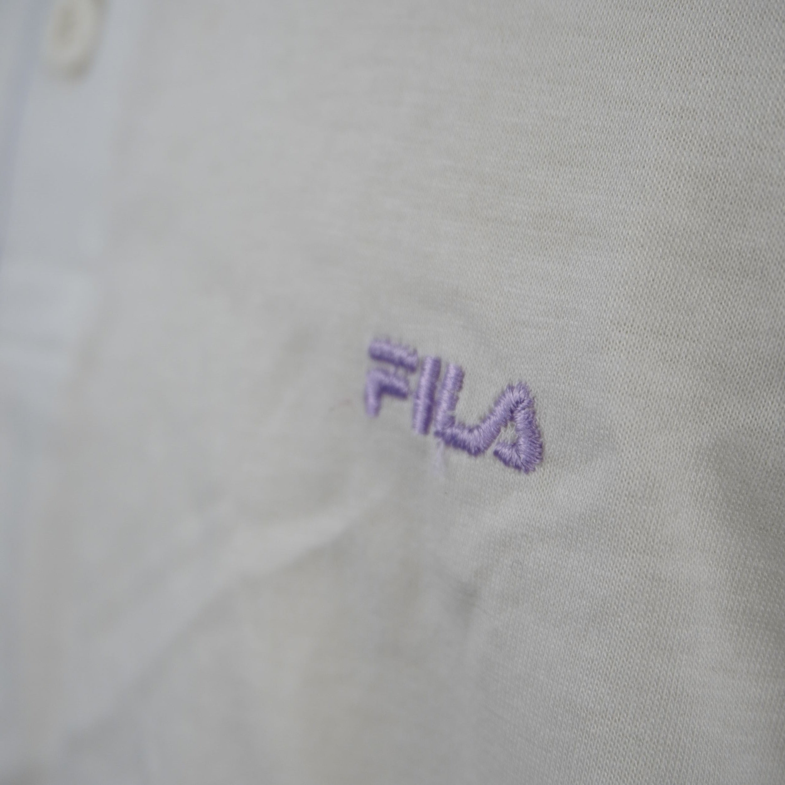 Macro sul ricamo del logo FILA in filo lilla pastello posizionato sul petto color panna di una polo vintage originale.