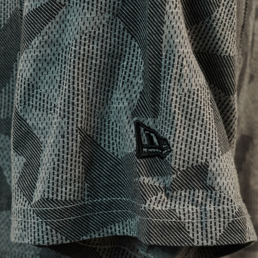 Inquadratura ravvicinata del bordo della manica corta con il piccolo logo iconico di New Era ricamato in filo nero su sfondo mimetico grigio.