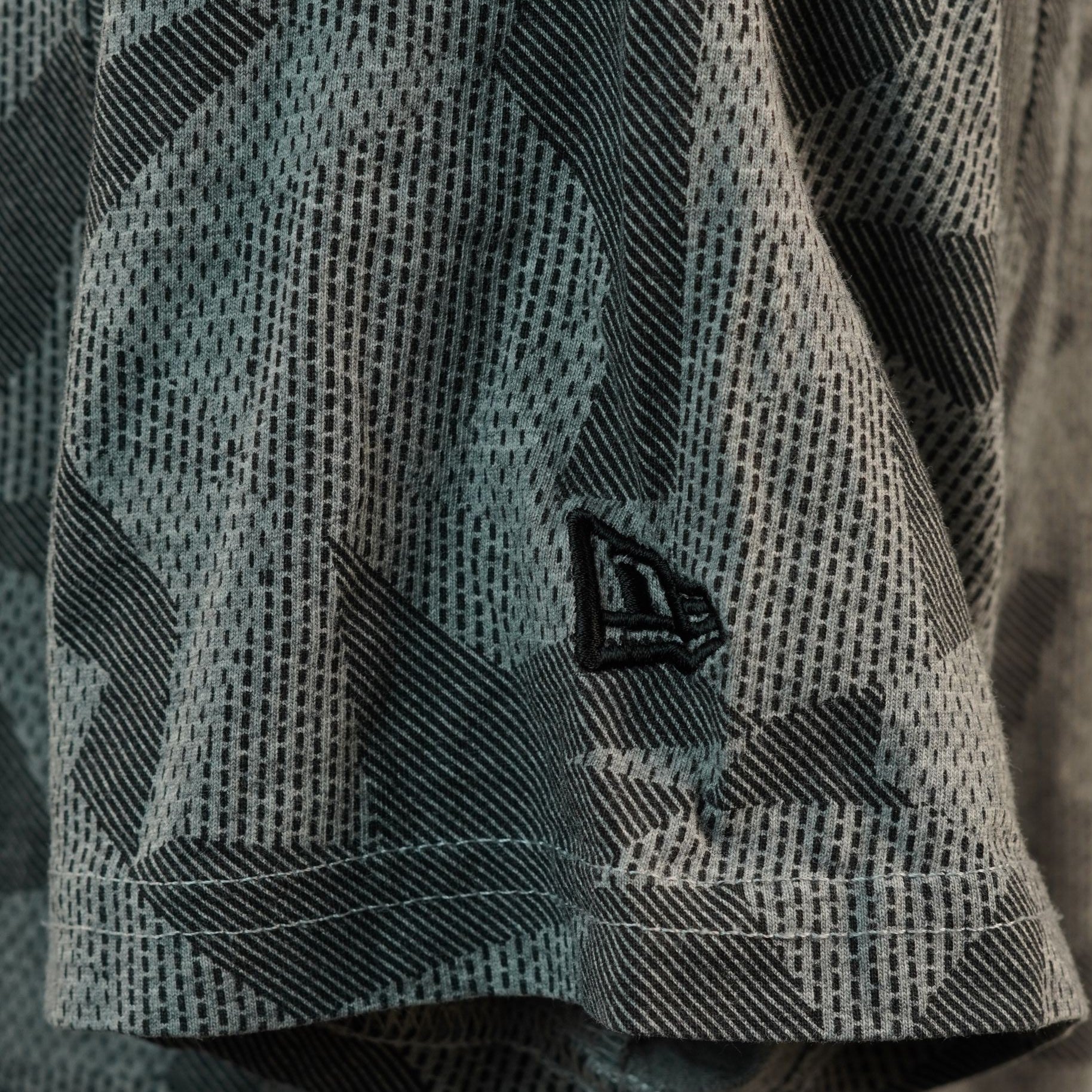 Inquadratura ravvicinata del bordo della manica corta con il piccolo logo iconico di New Era ricamato in filo nero su sfondo mimetico grigio.
