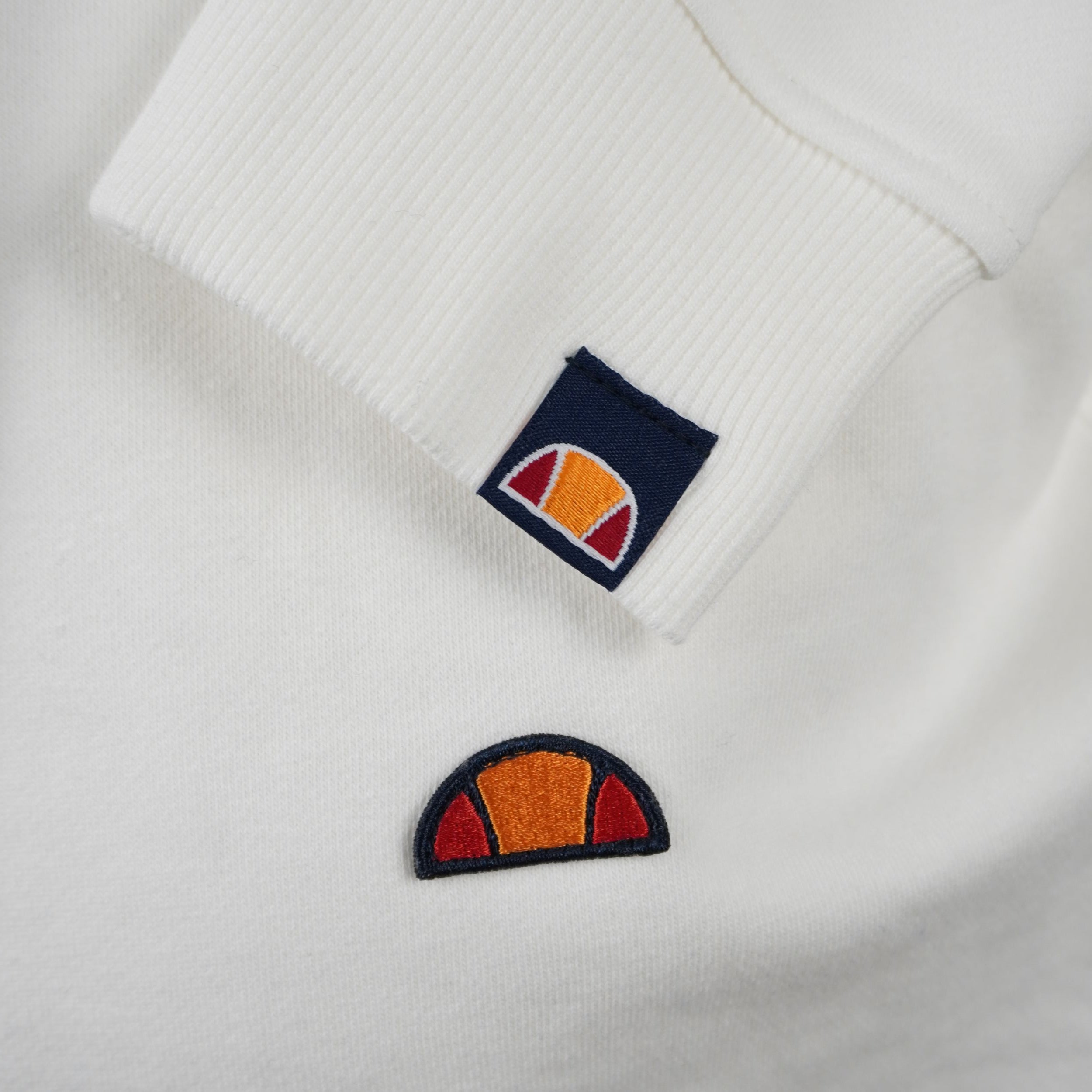 Primo piano ravvicinato del logo a semi-palla ellesse ricamato sul petto e del dettaglio sul polsino.