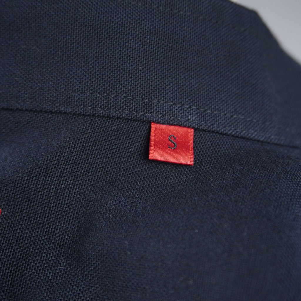 Dettaglio della piccola etichetta rossa con logo HUGO cucita sulla cucitura laterale della camicia nera.