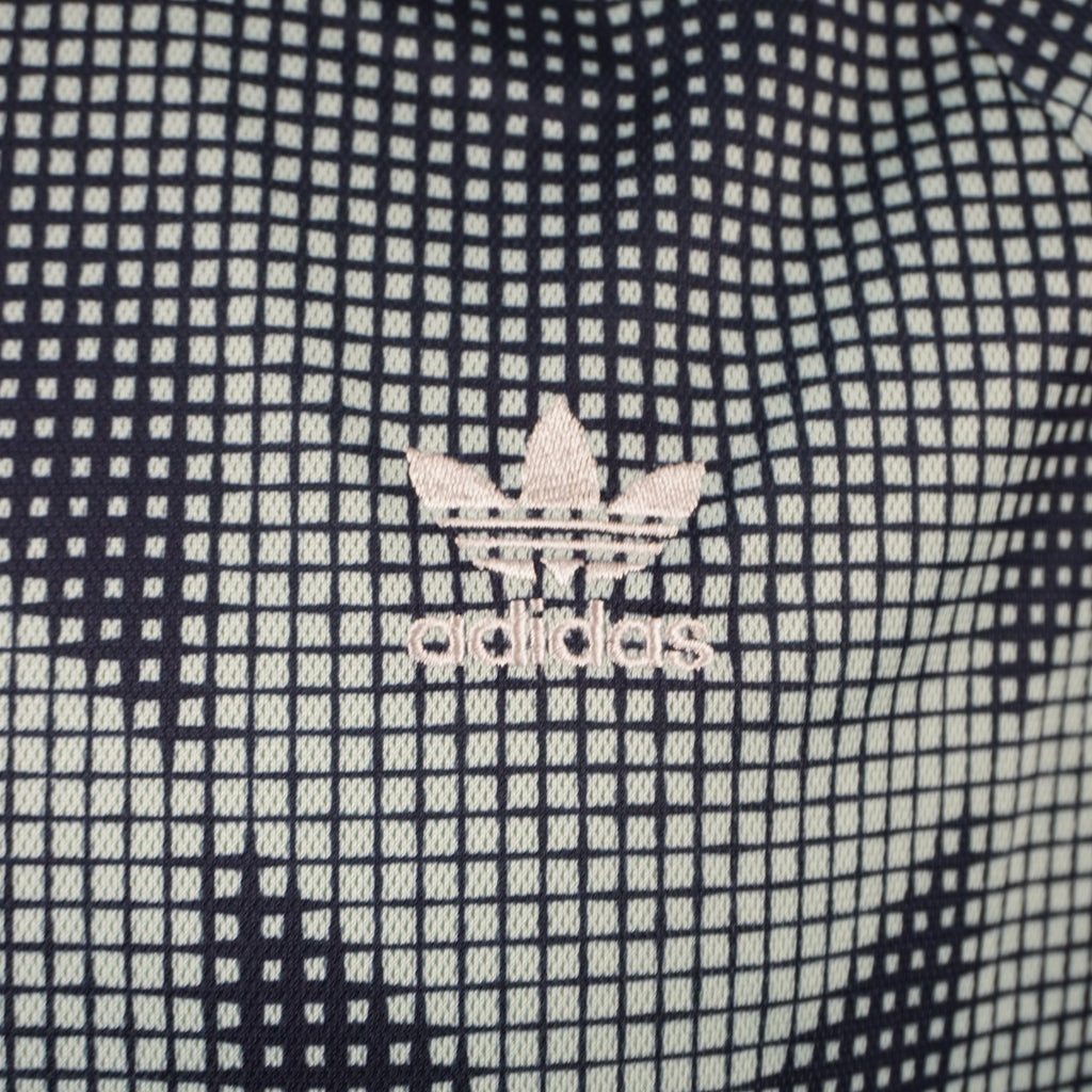 Primo piano del classico logo a trifoglio adidas ricamato in bianco sulla trama quadrettata a scacchi della t-shirt.