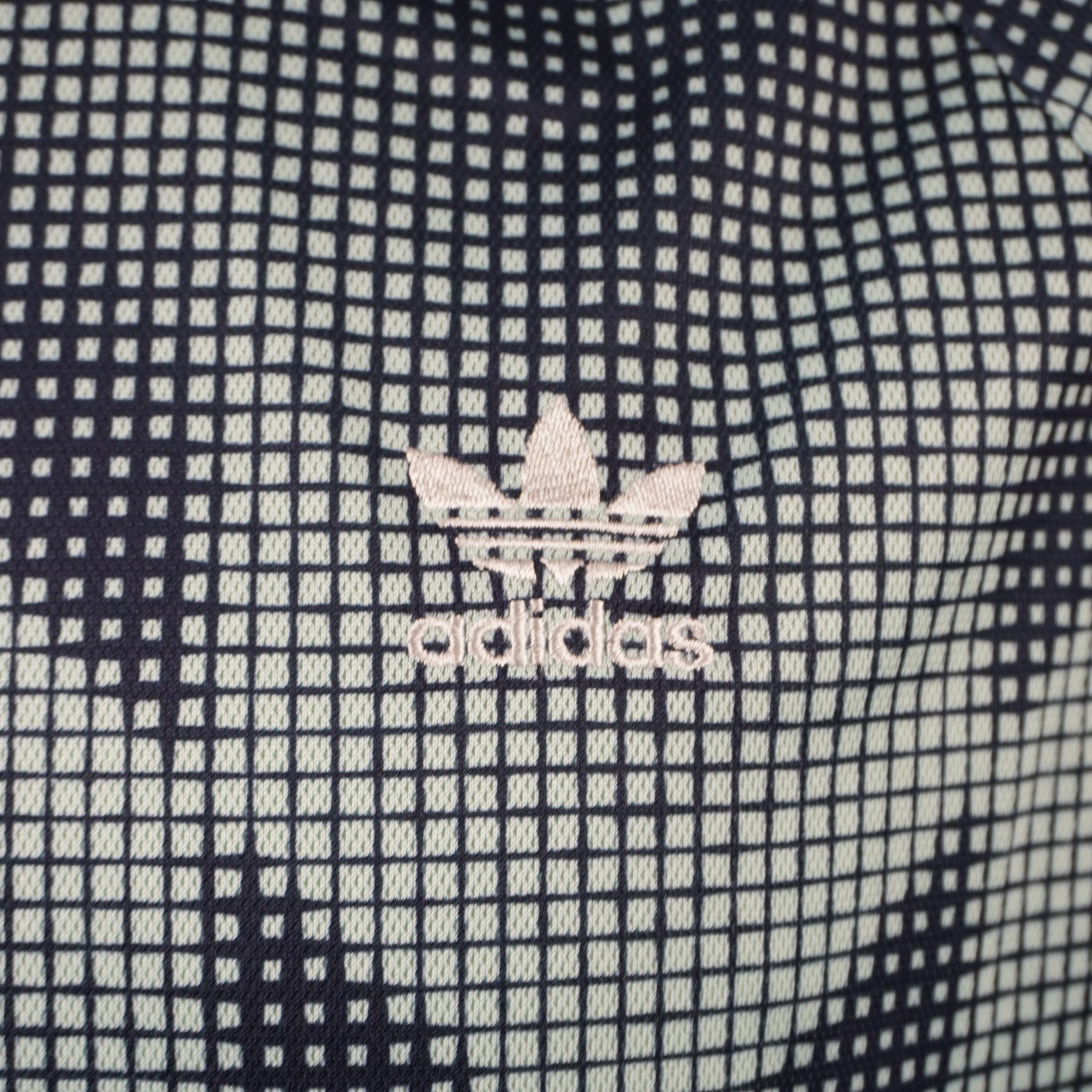 Primo piano del classico logo a trifoglio adidas ricamato in bianco sulla trama quadrettata a scacchi della t-shirt.