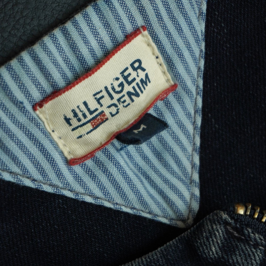  Inquadratura ravvicinata dell'etichetta interna rettangolare con la scritta ricamata Hilfiger Denim su sfondo chiaro.
