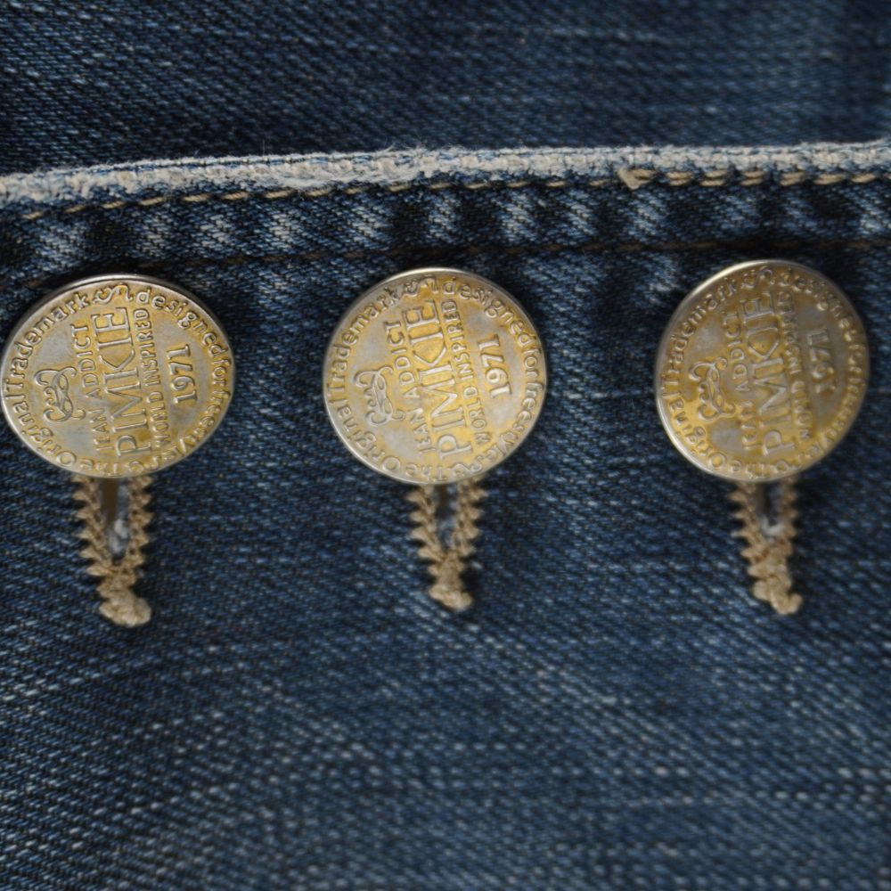 Primo piano dei tre maxi bottoni metallici a chiodo in stile bronzo antico incisi con il logo d'archivio Pimkie Jean Addict.