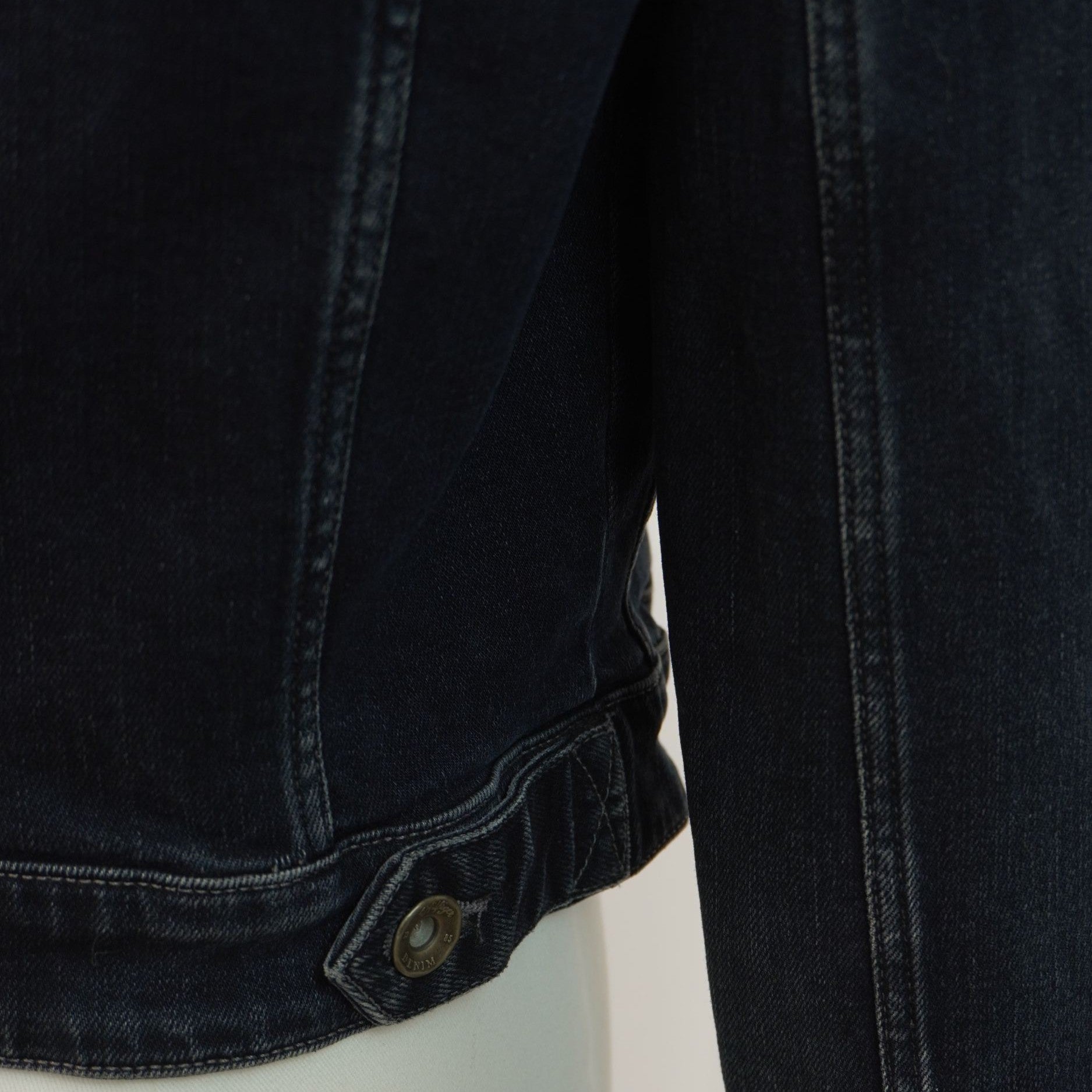 Vista ravvicinata dell'orlo inferiore della giacca di jeans con focus sul bottone metallico personalizzato applicato sulla linguetta laterale.
