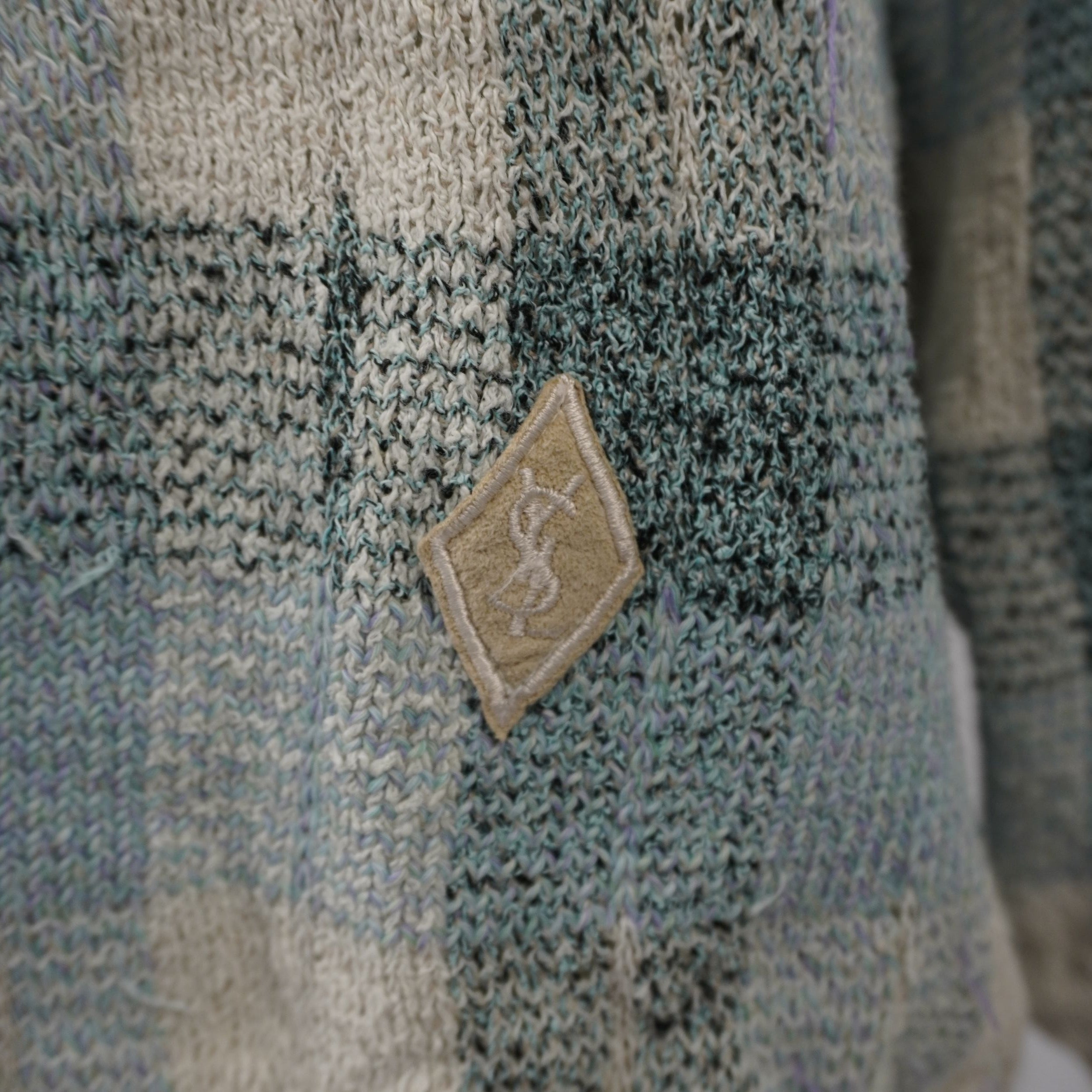 Dettaglio ravvicinato della patch a diamante marrone con il monogramma YSL ricamato vicino all'orlo del maglione.