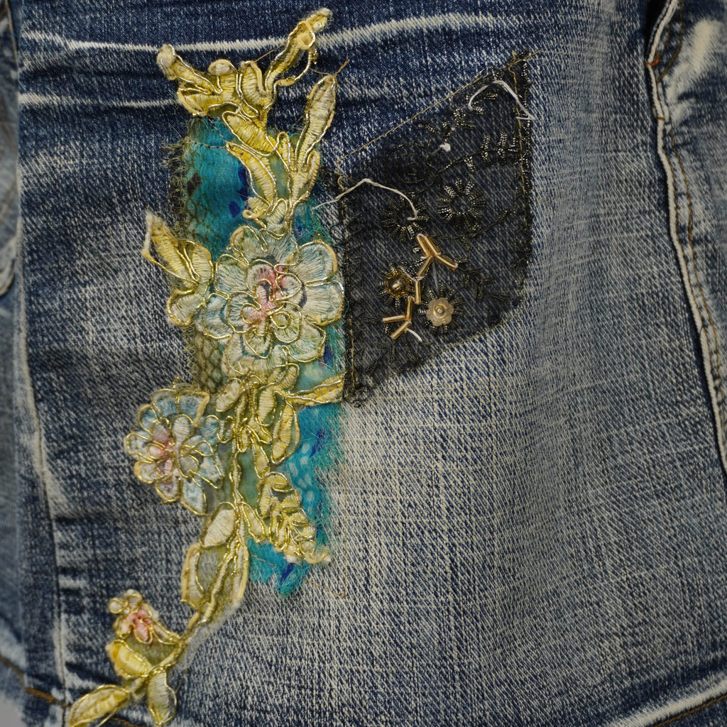 Inquadratura ravvicinata del patchwork boho-chic con pizzo, tulle azzurro e fiori ricamati sulla minigonna in denim.