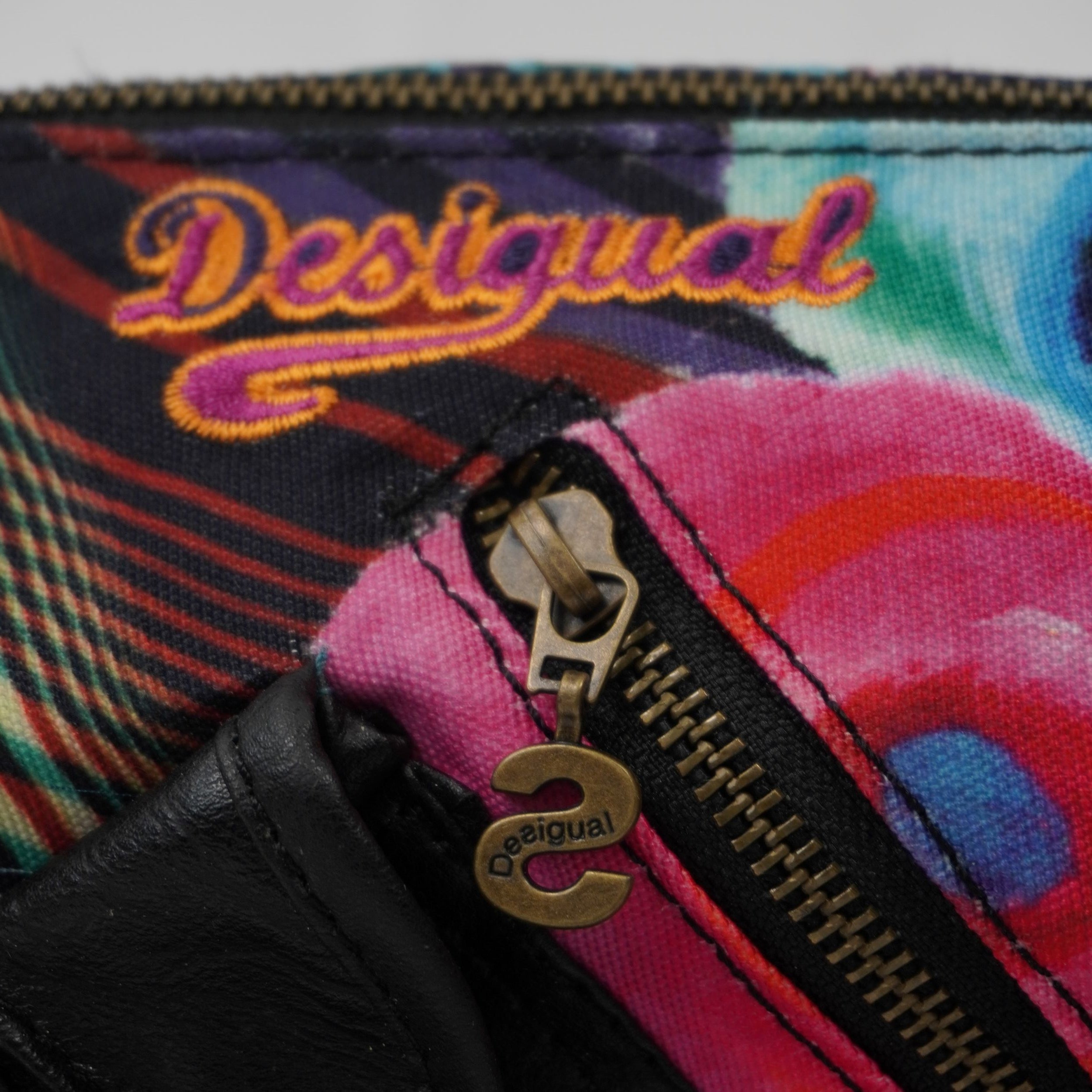 Primo piano della parte superiore della borsa che mostra il logo Desigual ricamato in filo arancione e una cerniera con cursore decorativo.