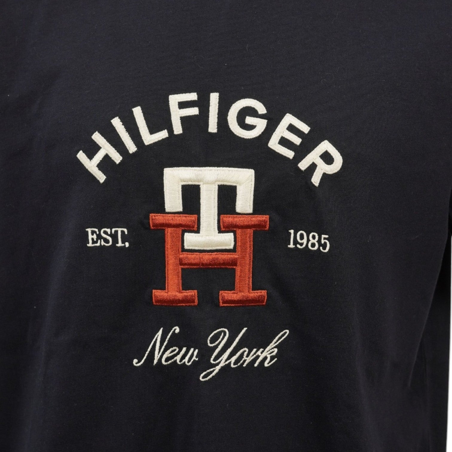 Primo piano del logo ricamato arancione TH e della scritta Hilfiger New York EST. 1985 sul petto.