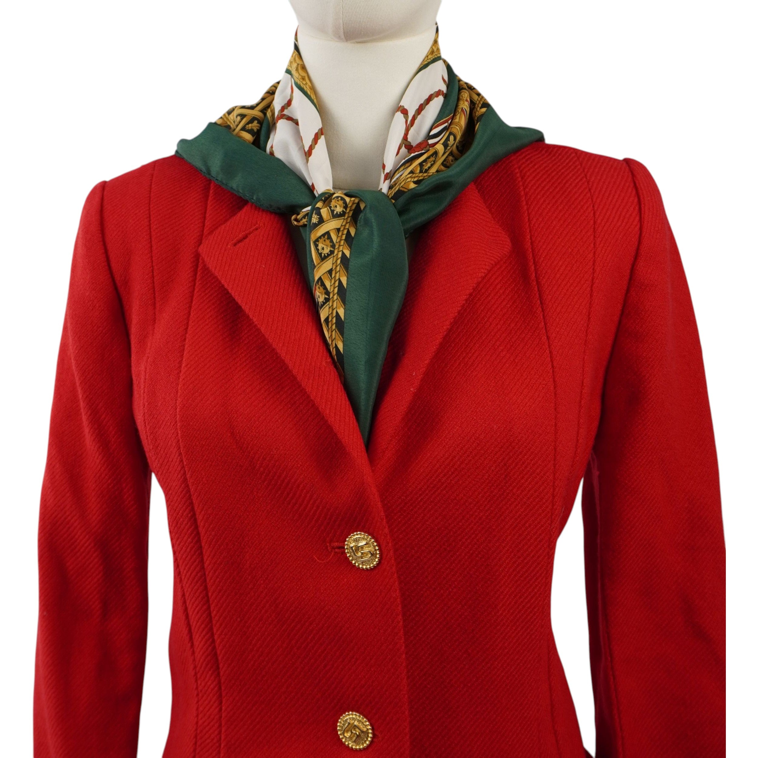 Primo piano della parte superiore della giacca rossa, con colletto a revers e un foulard in seta fantasia verde e oro elegantemente annodato sul décolleté.