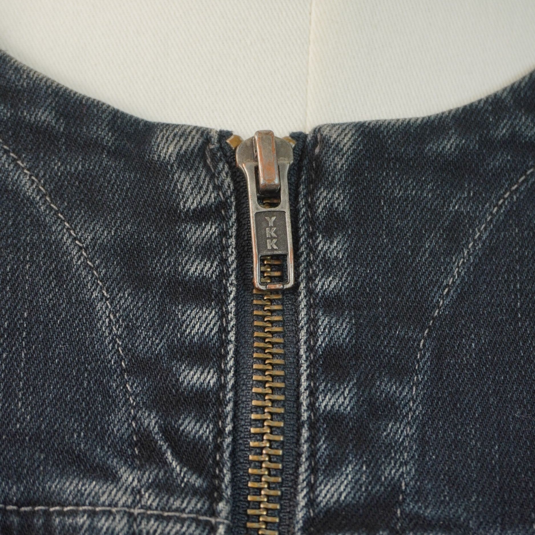 Primo piano dello scollo rotondo senza colletto della giacca, che evidenzia la chiusura lampo in metallo e le impunture sul denim.
