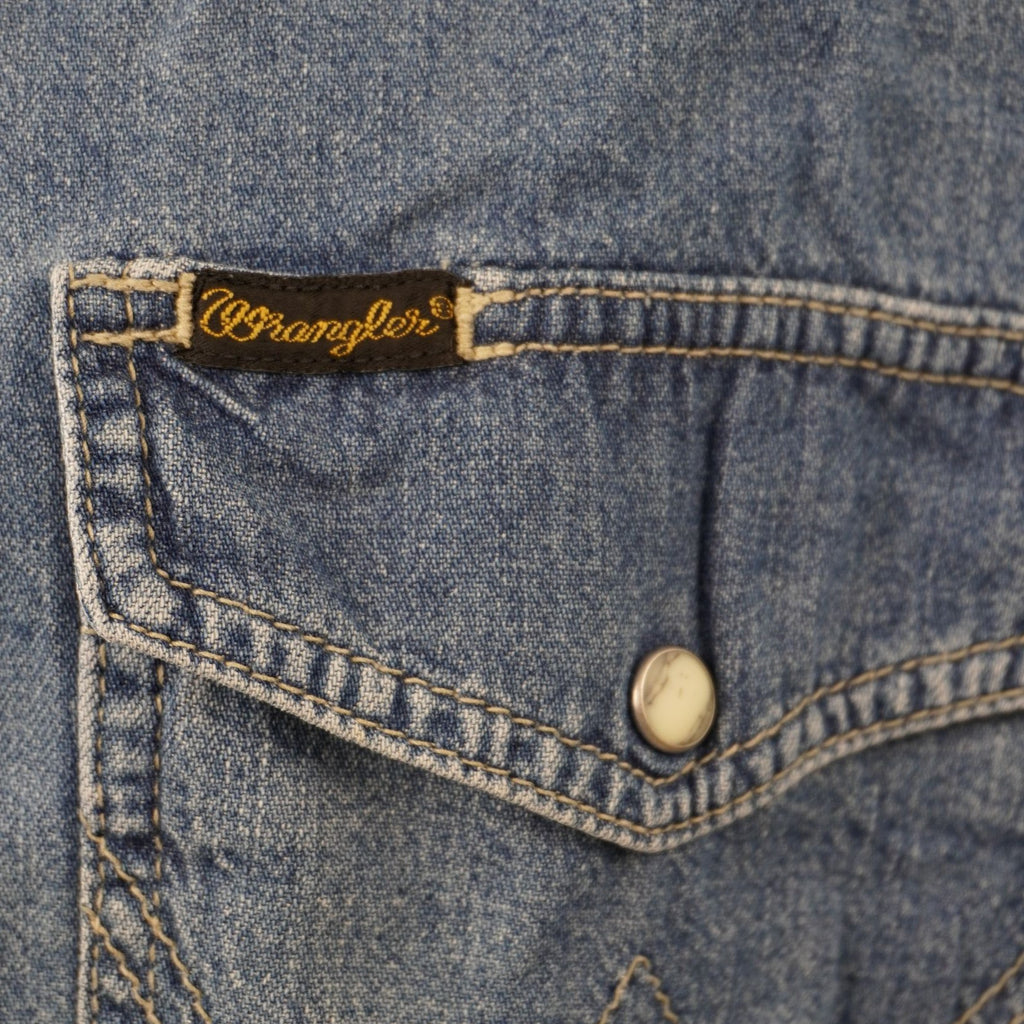 Inquadratura ravvicinata della tasca sinistra sul petto con patta sagomata, bottone in madreperla e piccola etichetta marrone con logo Wrangler.