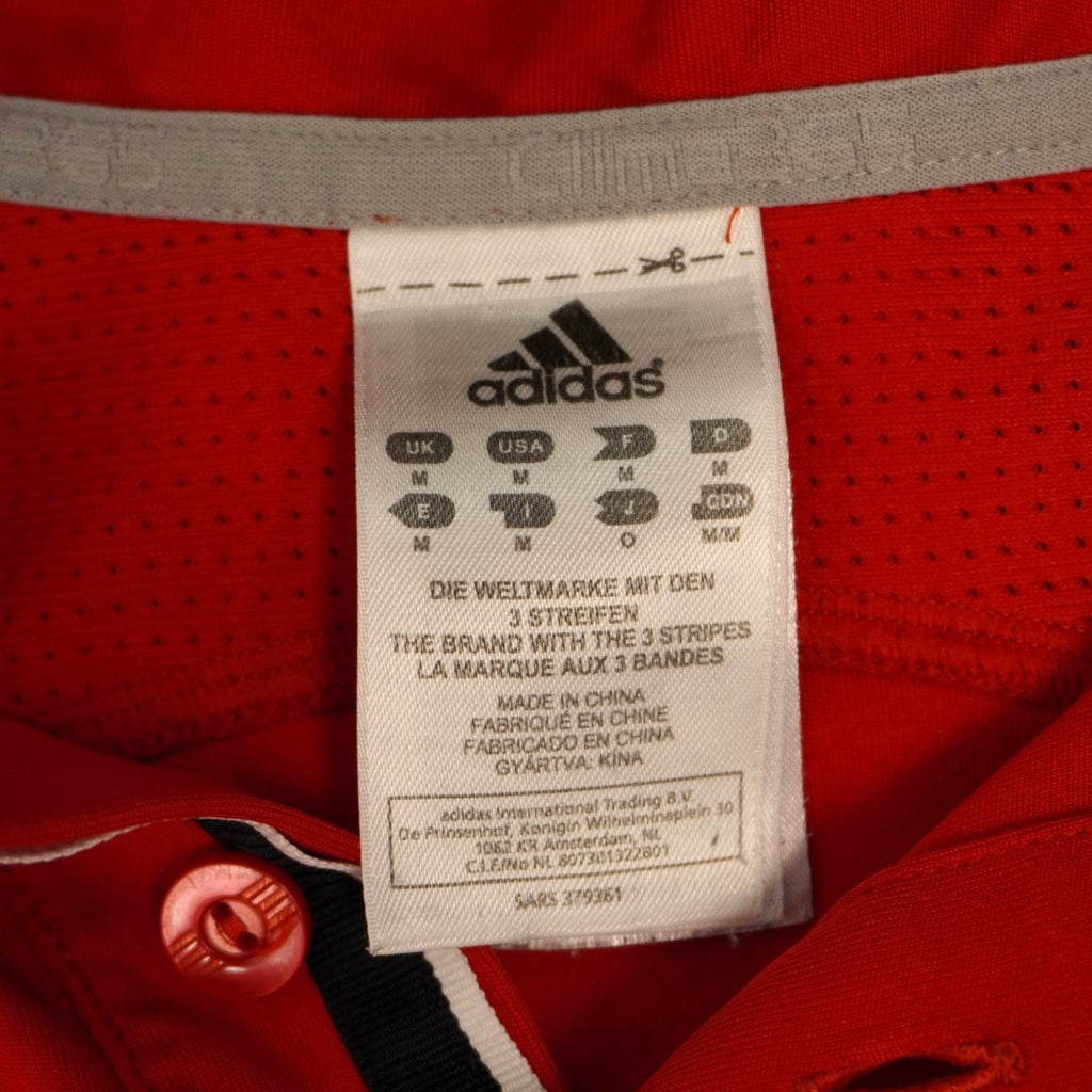 Etichetta interna cucita sul colletto della polo rossa Adidas Barricade che mostra il logo del brand e la taglia M.