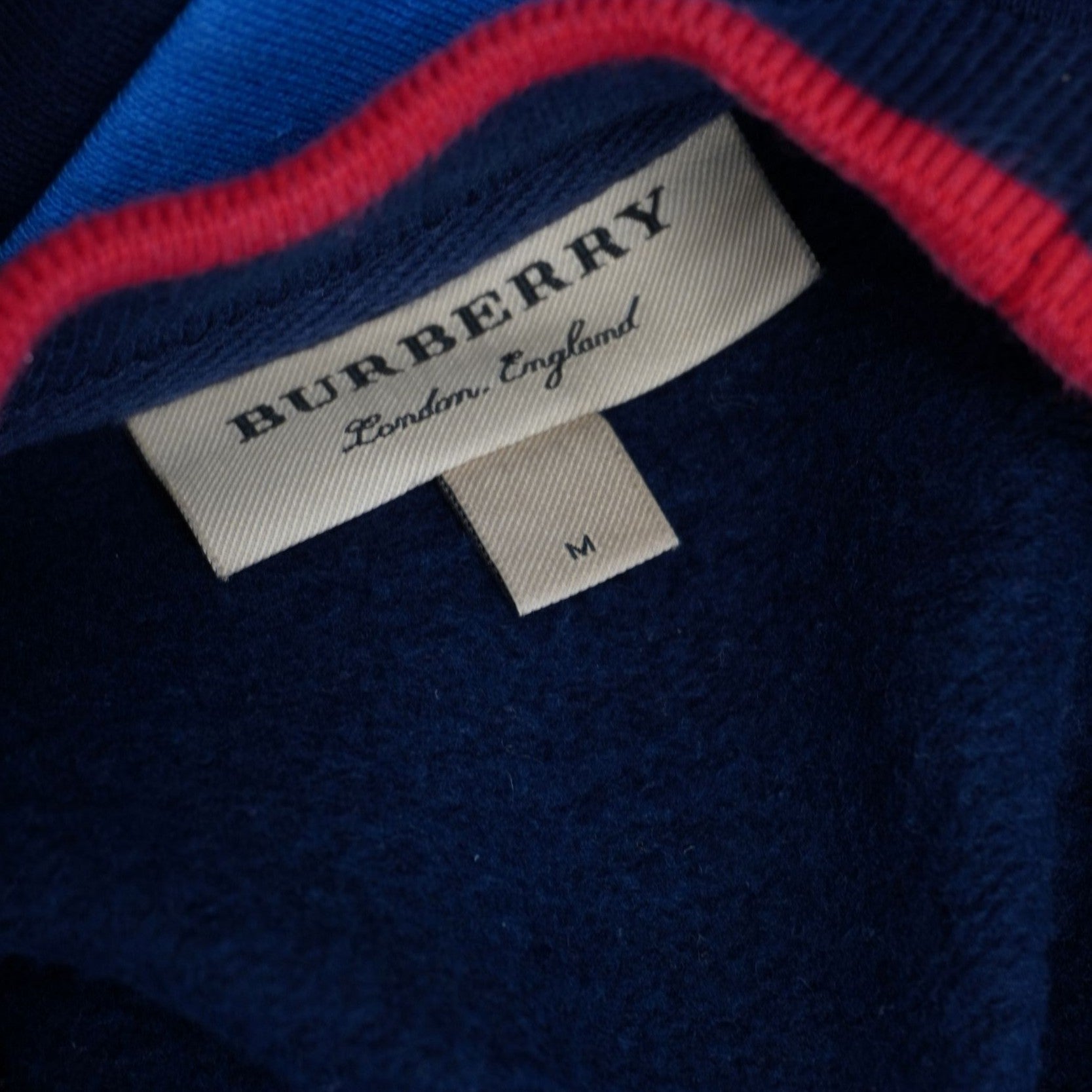 Dettaglio dell'etichetta rettangolare beige con il logo Burberry London England cucita all'interno del colletto sopra il cartellino della taglia M