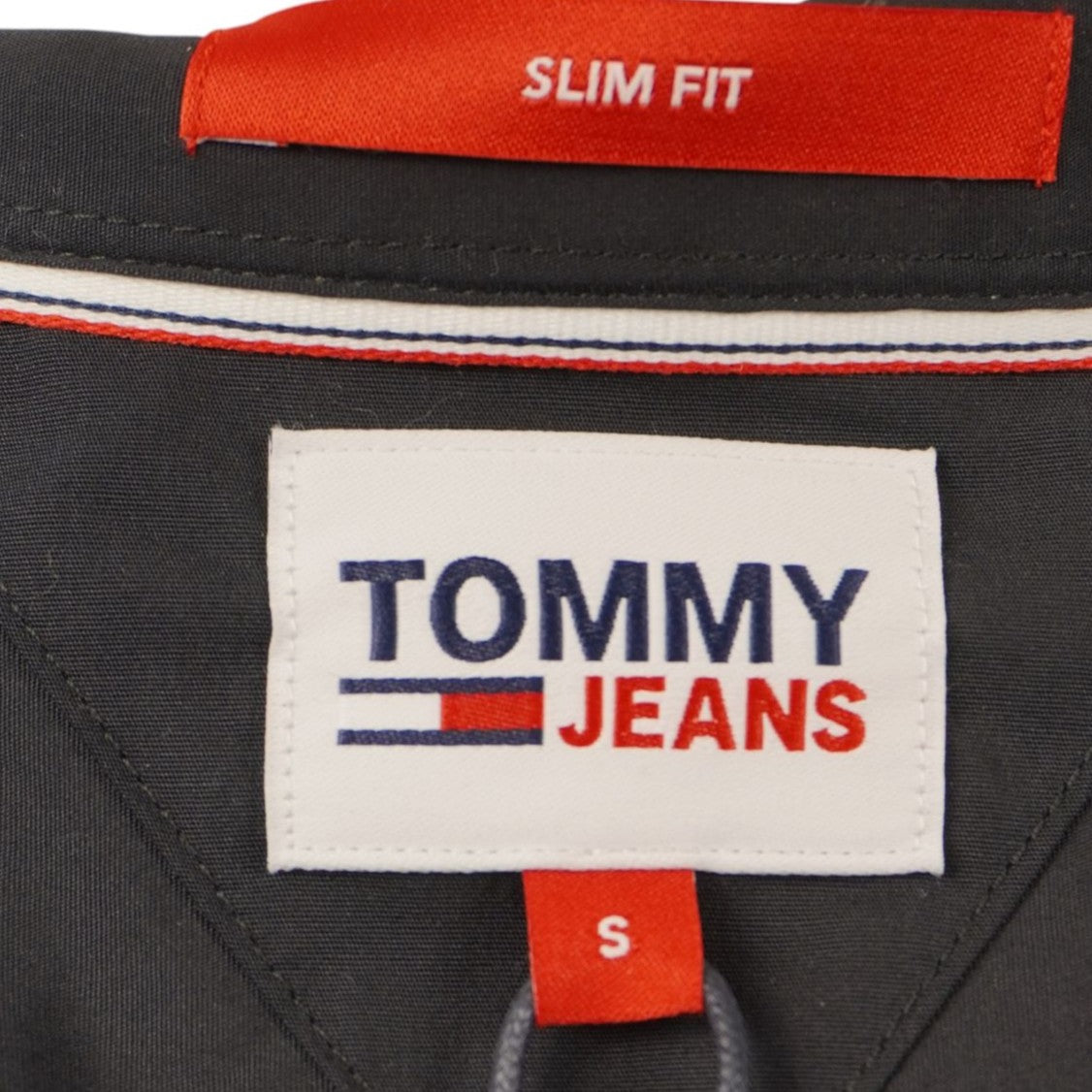 Dettaglio del logo Tommy Jeans rettangolare con profilo rosso e indicazione della taglia S ricamati internamente sotto il colletto.