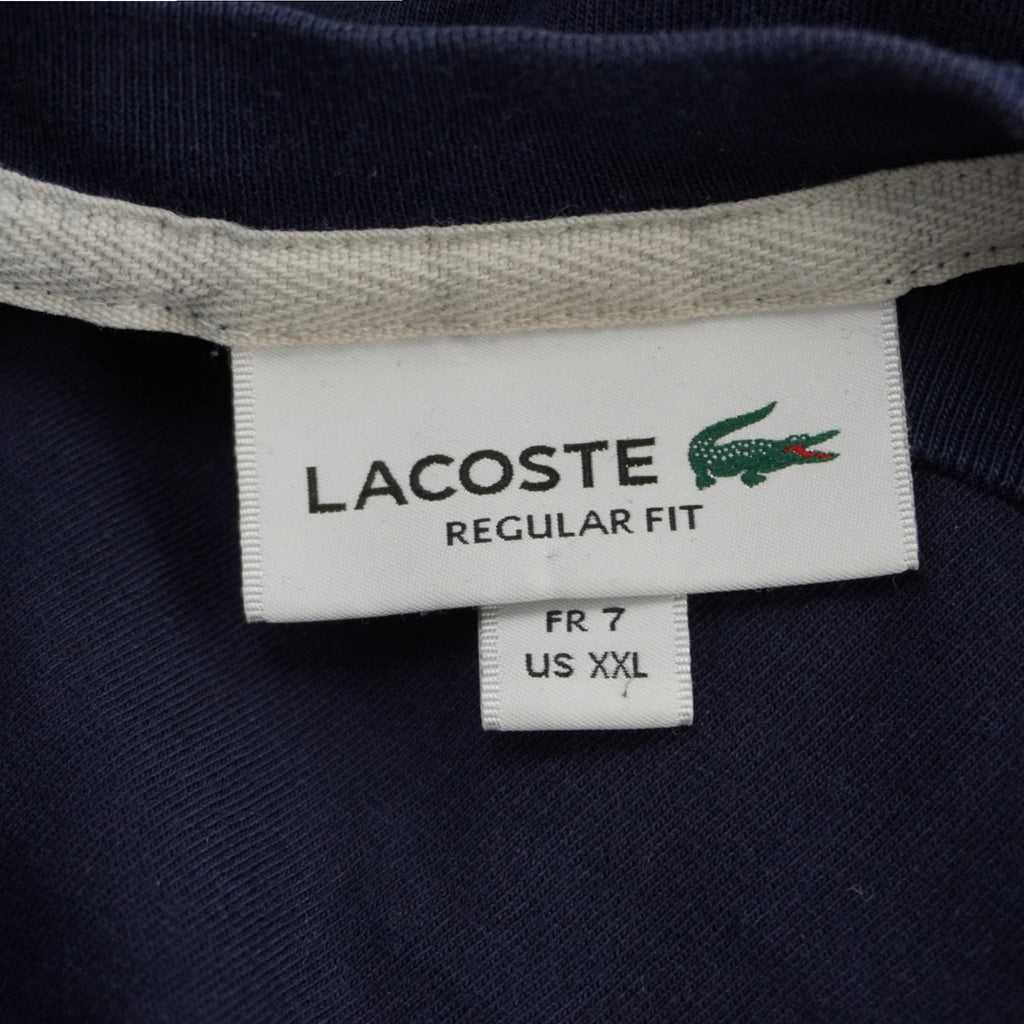 Primo piano dell'etichetta bianca interna sul colletto della t-shirt Lacoste con logo del coccodrillo verde, scritta Regular Fit e indicazione della taglia FR 7 e US XXL.