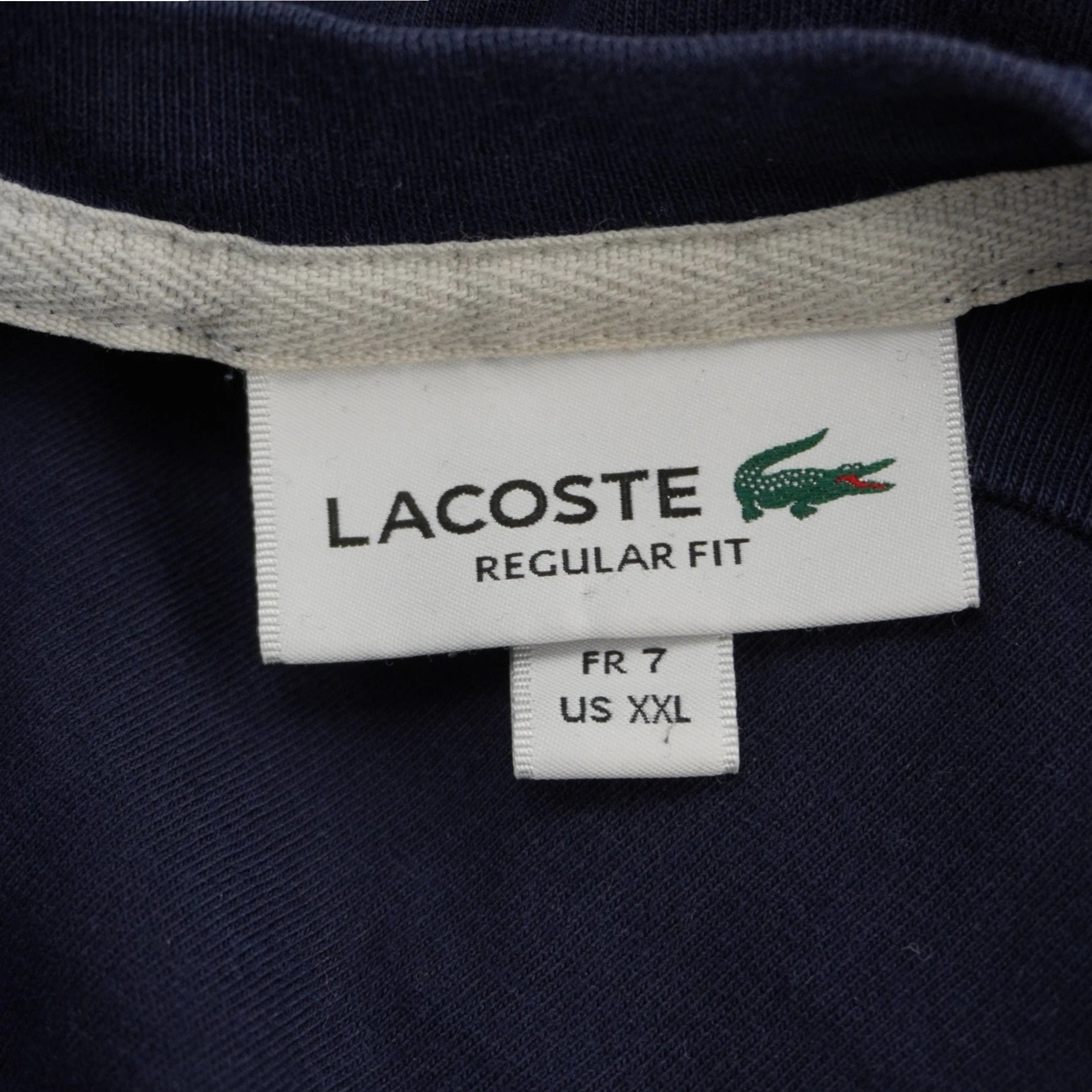 Primo piano dell'etichetta bianca interna sul colletto della t-shirt Lacoste con logo del coccodrillo verde, scritta Regular Fit e indicazione della taglia FR 7 e US XXL.