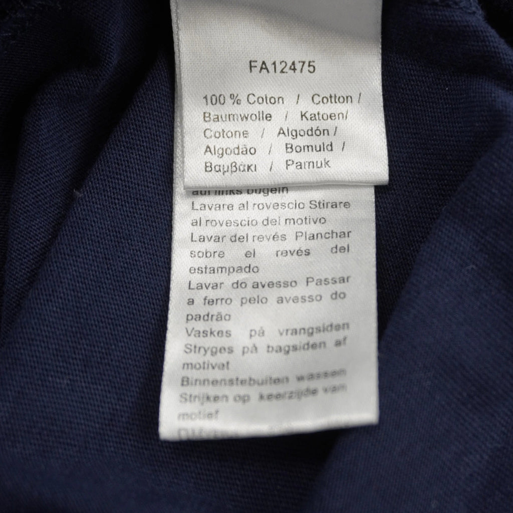 Dettaglio dell'etichetta interna di composizione della t-shirt Lacoste, che riporta il codice FA12475, la dicitura 100% cotone e le istruzioni di lavaggio in diverse lingue.