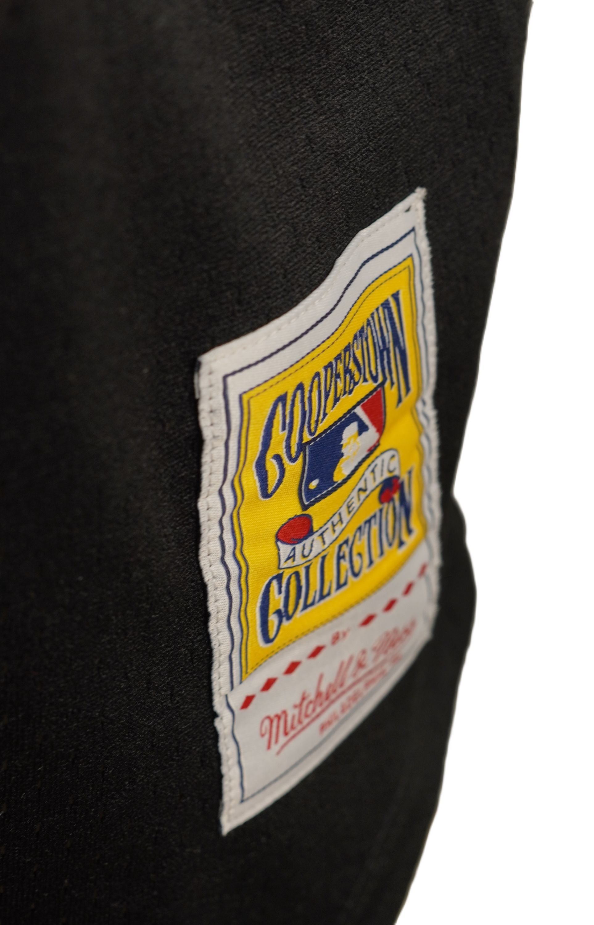 Dettaglio della patch rettangolare Cooperstown Collection firmata Mitchell & Ness cucita nell'orlo inferiore frontale della maglia.
