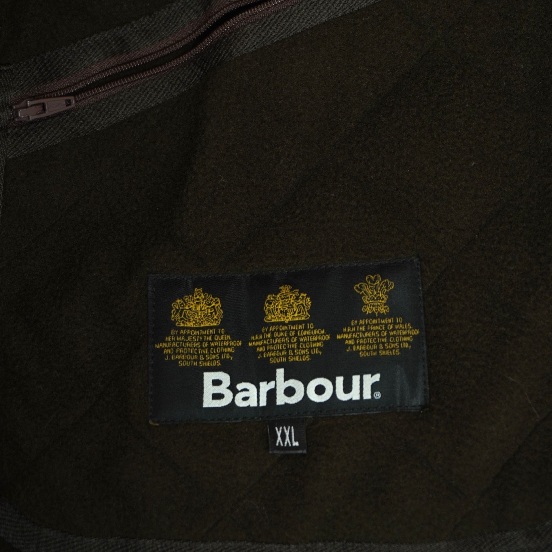 Primo piano dell'etichetta interna in tessuto nero con i tre loghi della casa reale britannica che attesta l'autenticità del capo Barbour.