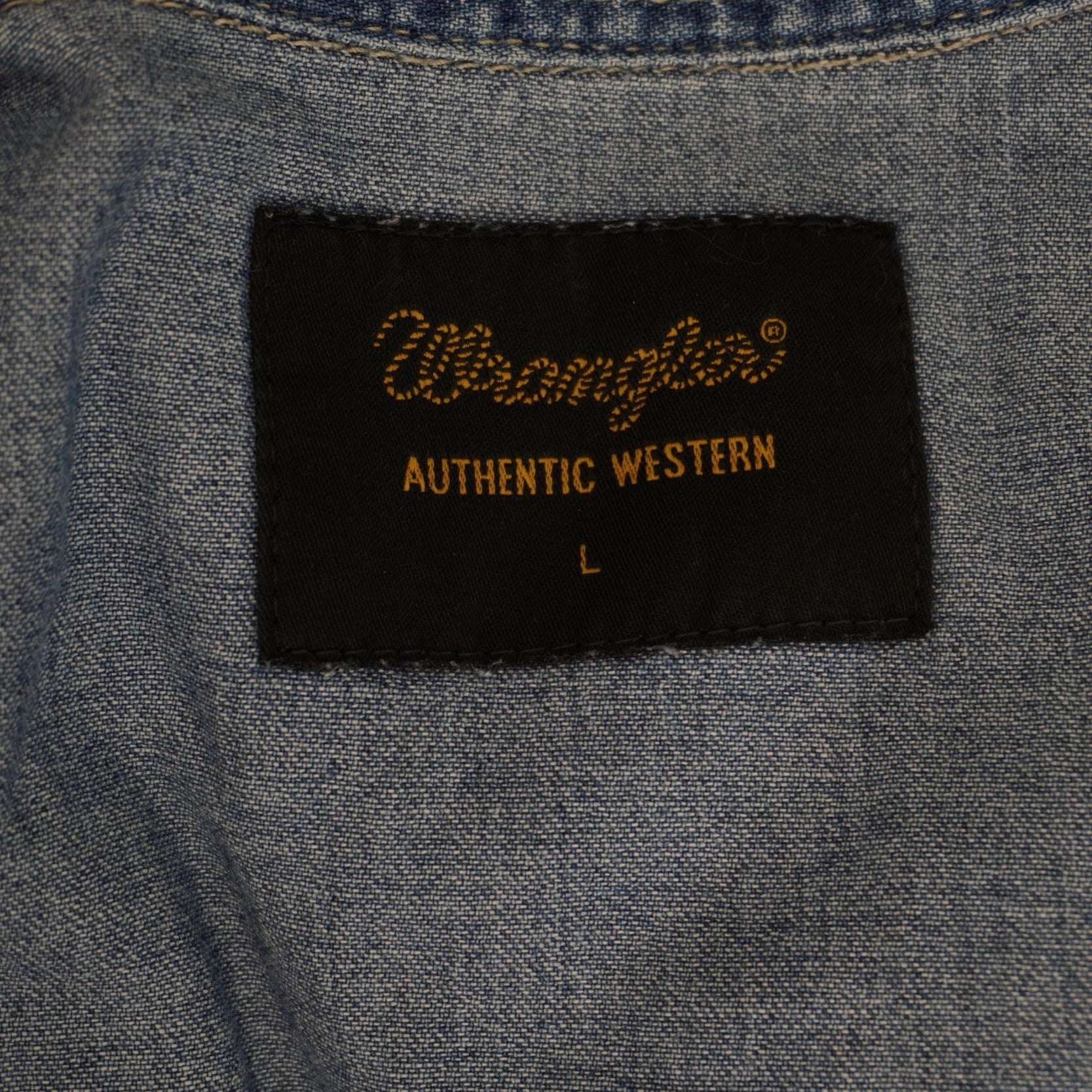 Inquadratura dell'etichetta rettangolare nera cucita sotto il colletto interno con la scritta dorata Wrangler Rugged Wear e la taglia L.