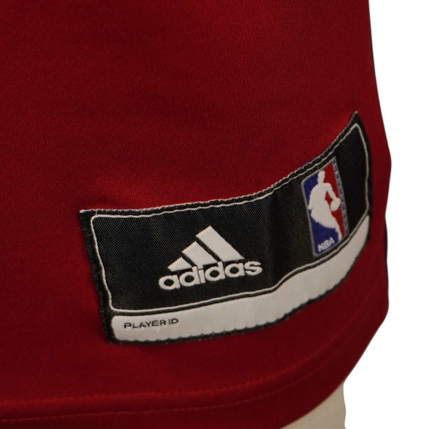 Dettaglio della targhetta rettangolare nera Adidas e NBA cucita nell'orlo inferiore frontale della canotta, con indicazione della taglia XS.
