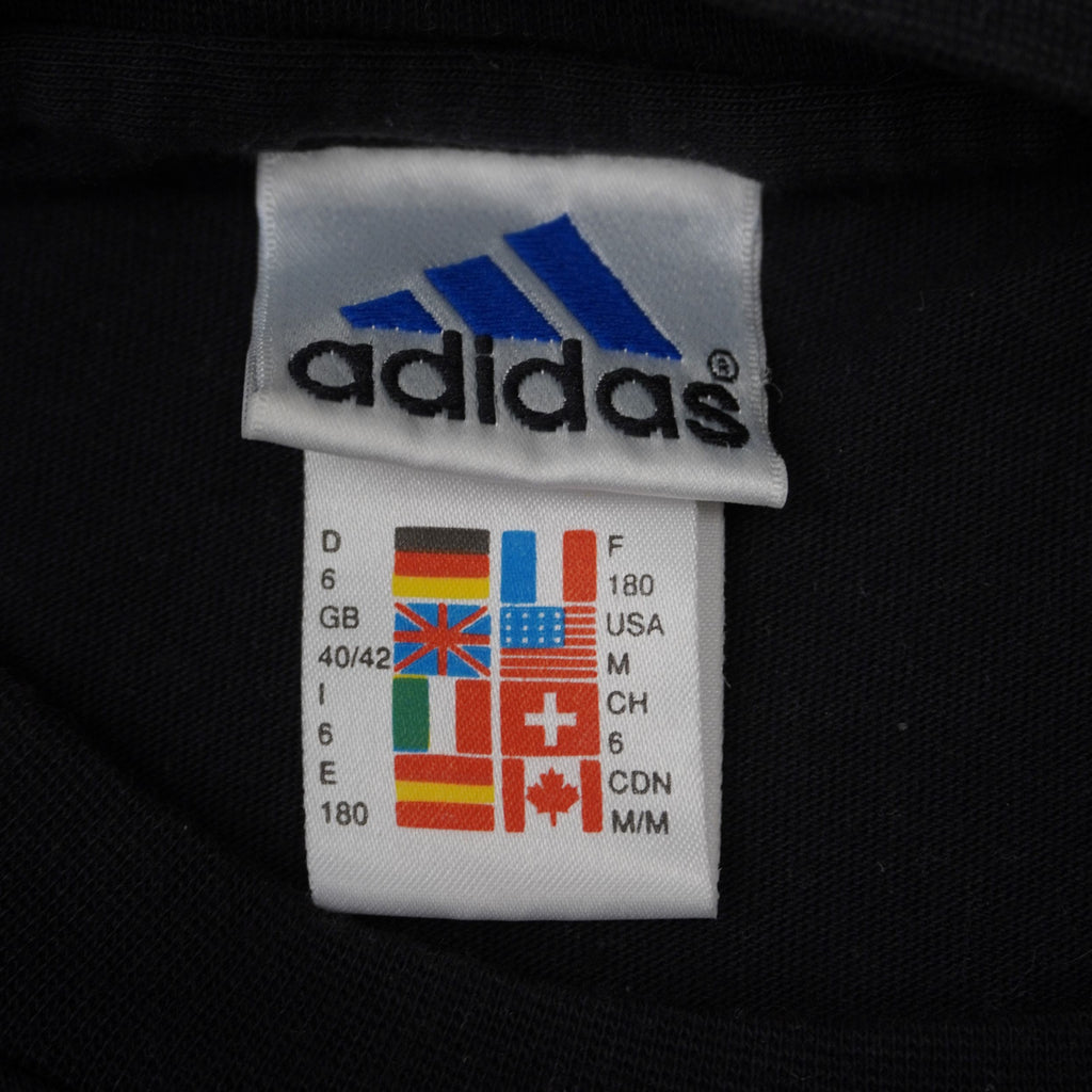 Etichetta interna Adidas vintage anni 90 con griglia multi-bandiera che mostra la taglia M per USA, Germania e Italia.