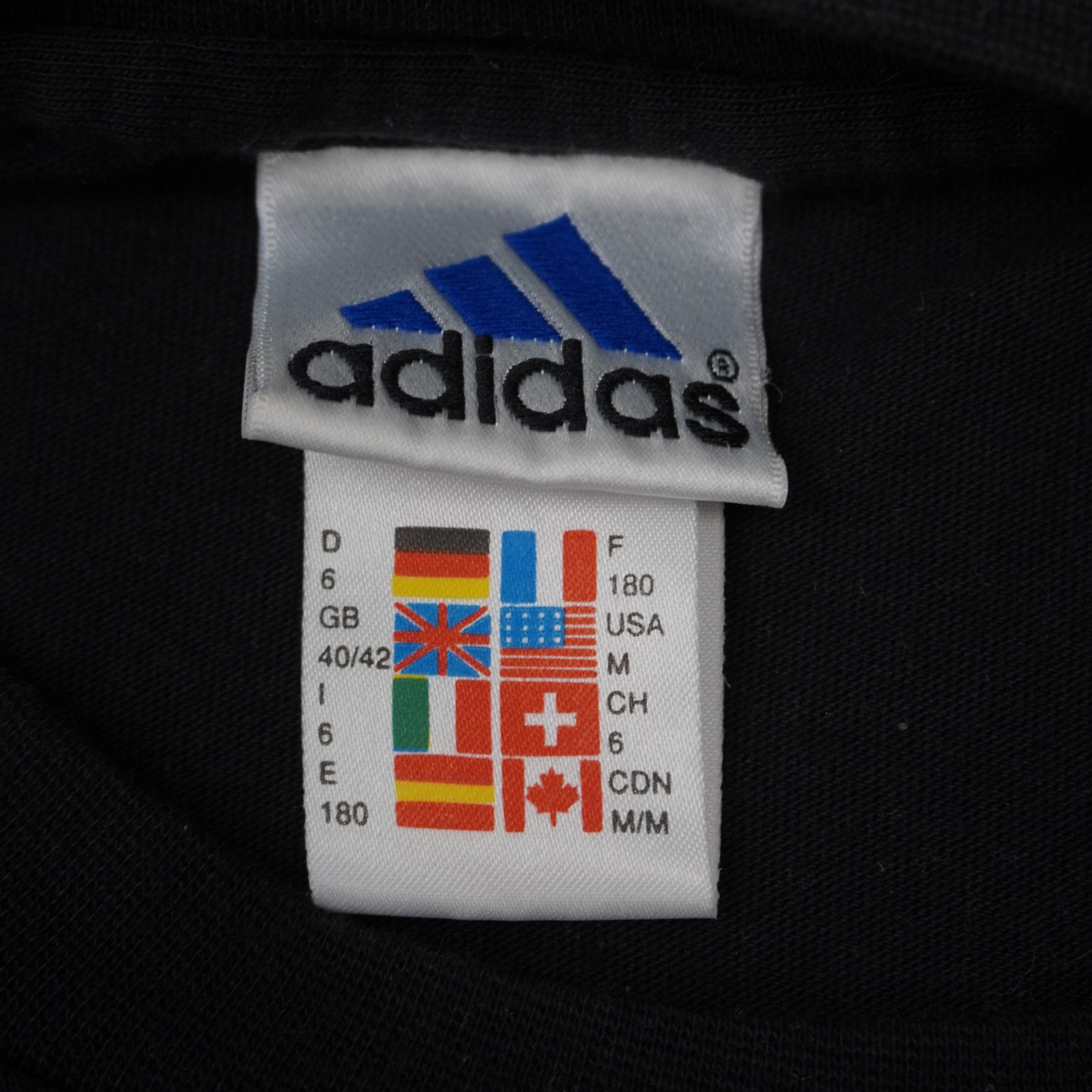 Etichetta interna Adidas vintage anni 90 con griglia multi-bandiera che mostra la taglia M per USA, Germania e Italia.