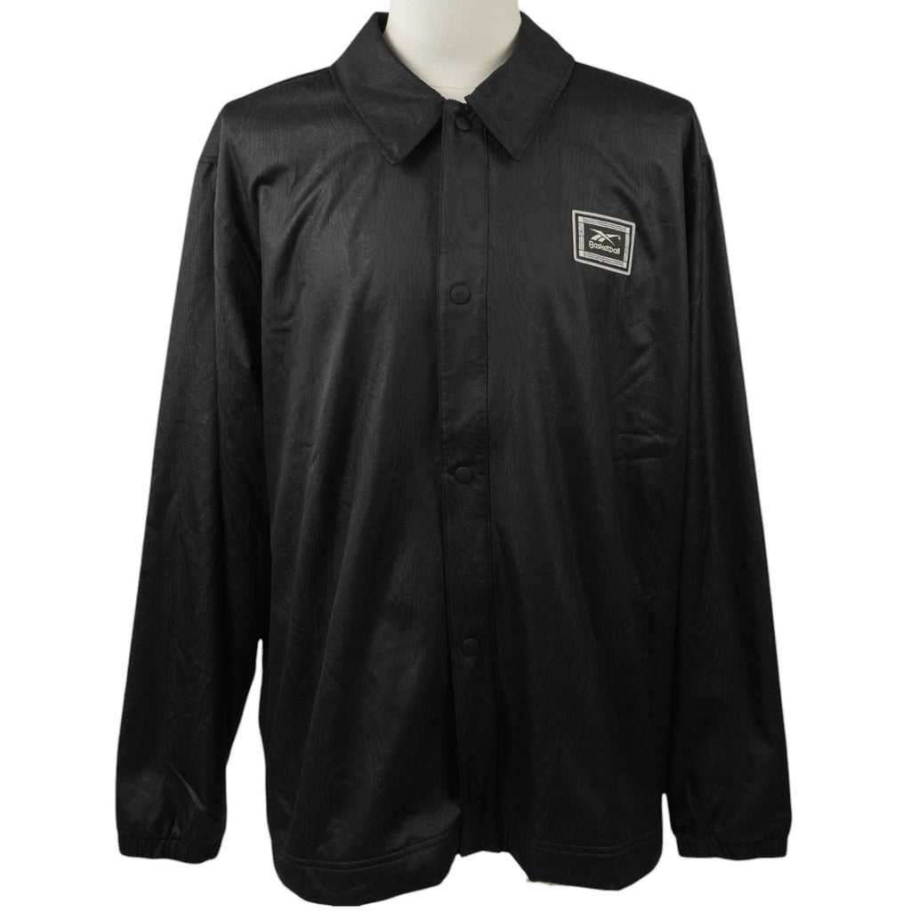 Vista frontale su manichino di una giacca sportiva Reebok nera modello Coach con colletto classico, chiusura con bottoni a pressione e patch quadrata ricamata sul petto.