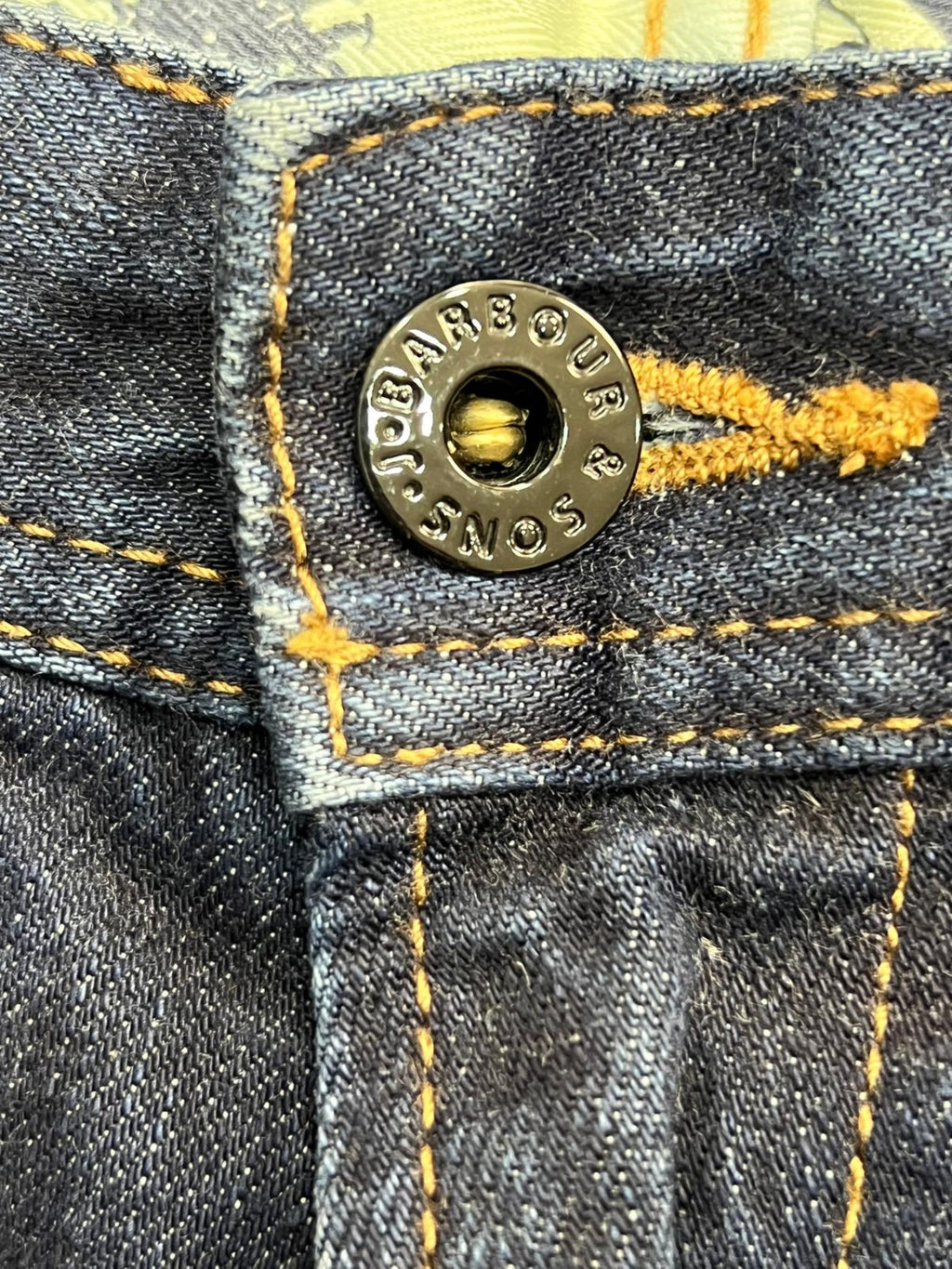 Macro del bottone principale in metallo brunito inciso con il logo Barbour International e asola profilata in filo giallo.