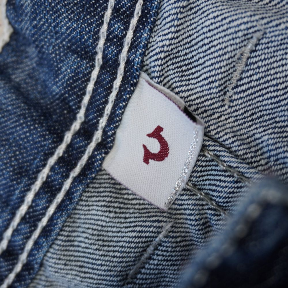 Primo piano dell'etichetta interna quadrata in tessuto bianco cucita sulla cintura dei jeans True Religion, con impresso il logo a ferro di cavallo rosso.