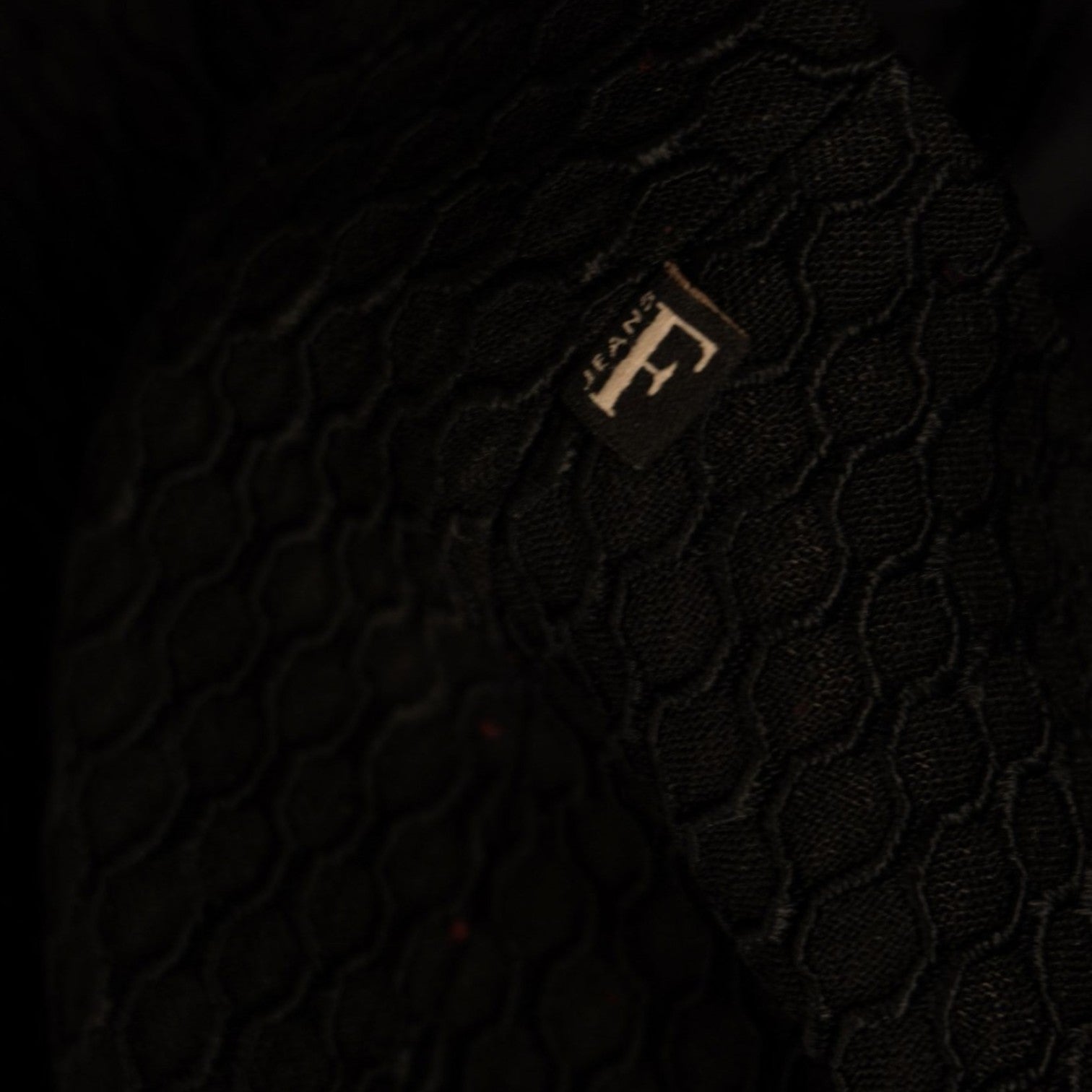 Dettaglio del ciondolo con logo a lettera F dorato sulla texture a nido d'ape.