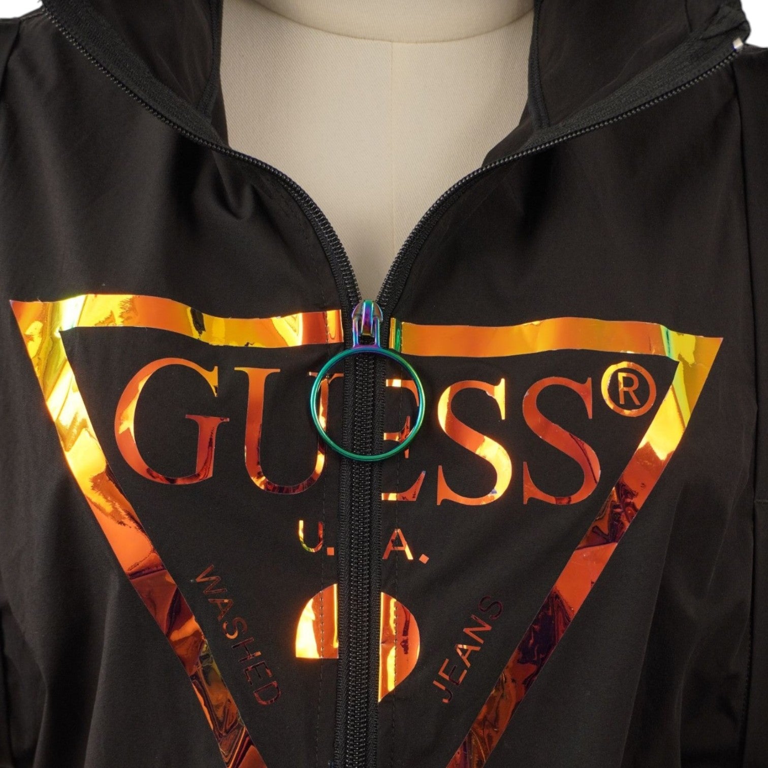 Dettaglio ravvicinato dell'iconico logo a triangolo Guess stampato in lamina metallizzata oro e bronzo.