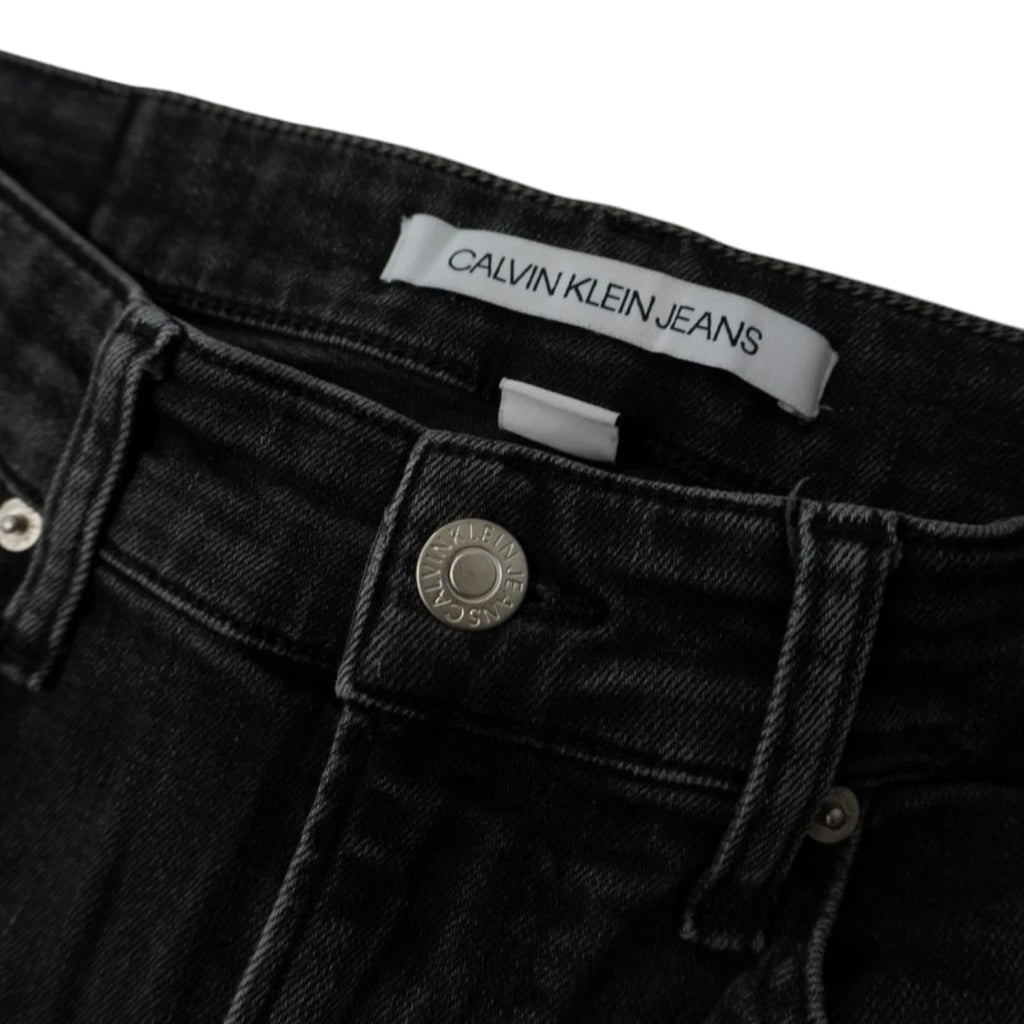 Dettaglio macro del bottone metallico inciso e dell'etichetta bianca interna Calvin Klein Jeans su denim grigio scuro