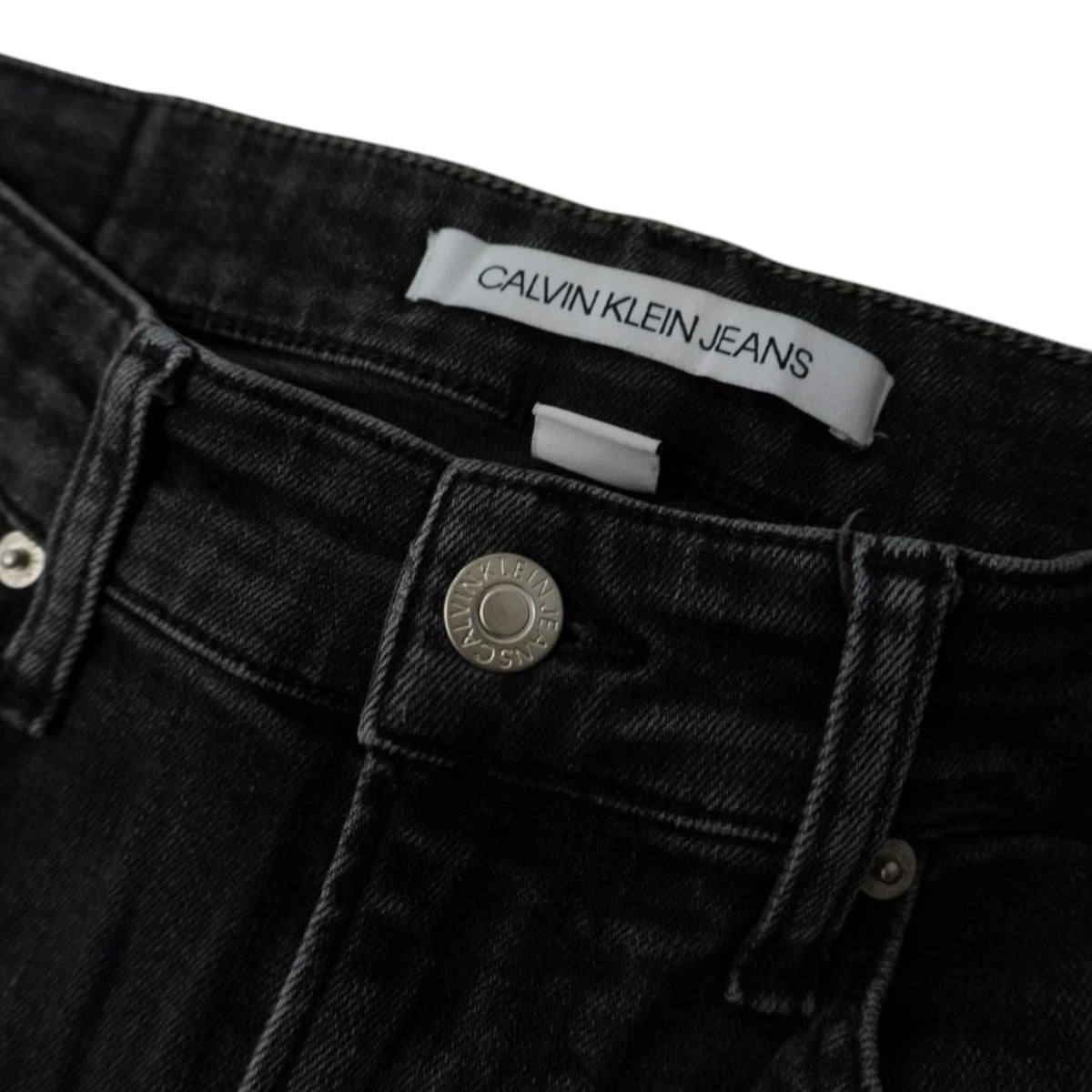 Dettaglio macro del bottone metallico inciso e dell'etichetta bianca interna Calvin Klein Jeans su denim grigio scuro